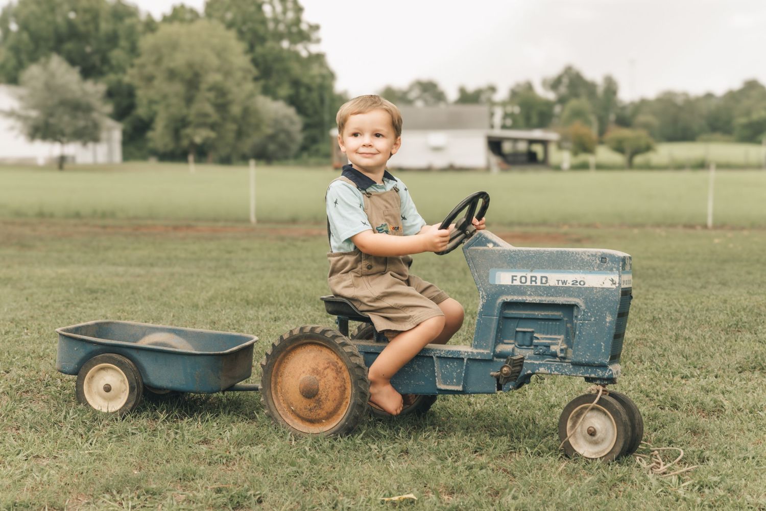 Toddler boy smiling while riding a vintage blue Ford pedal tractor with trailer on a green farm field.