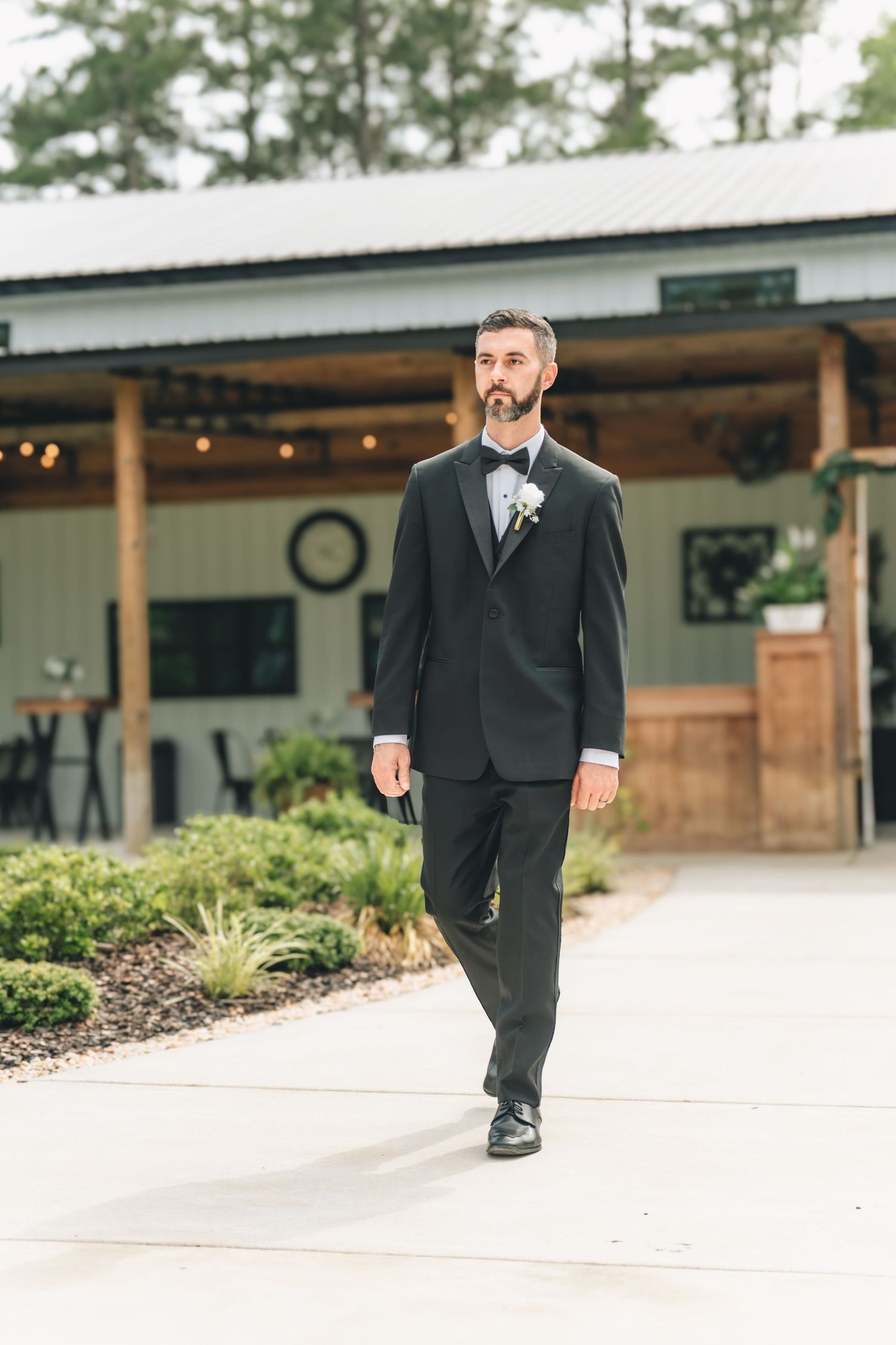 Groom in black tuxedo with boutonniere walks confidently outside rustic barn wedding venue.