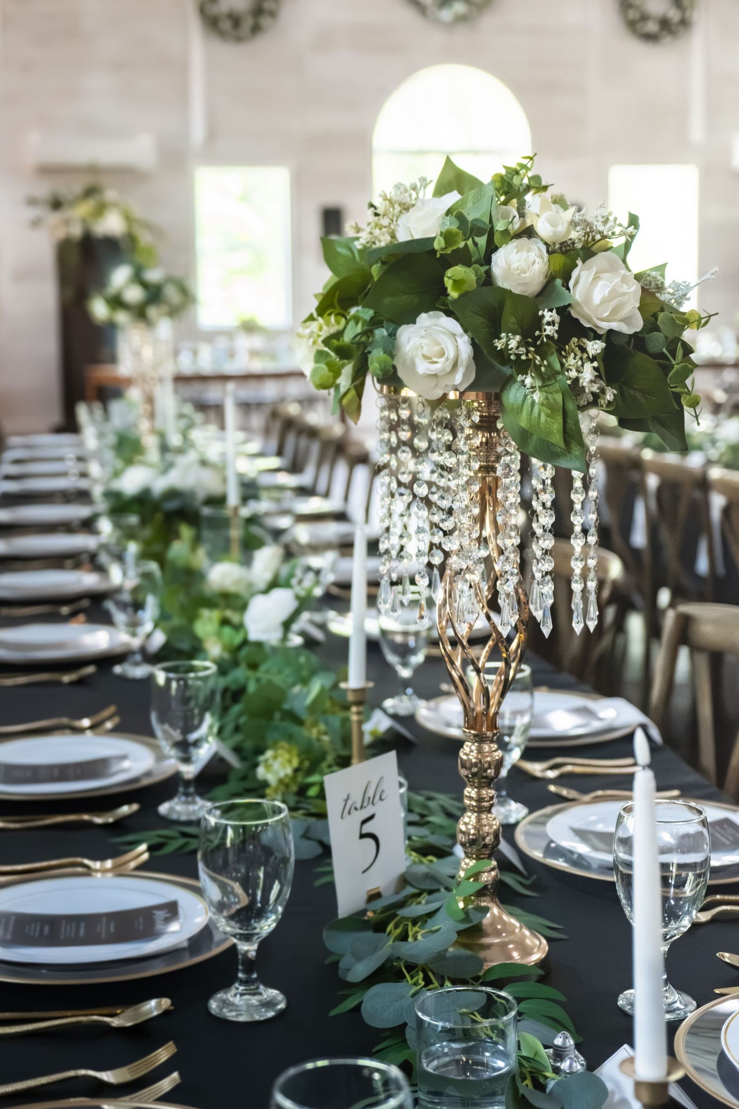 Elegant wedding reception table with gold candelabra, white roses, crystal drops, greenery, and black tablecloth with gold flatware.