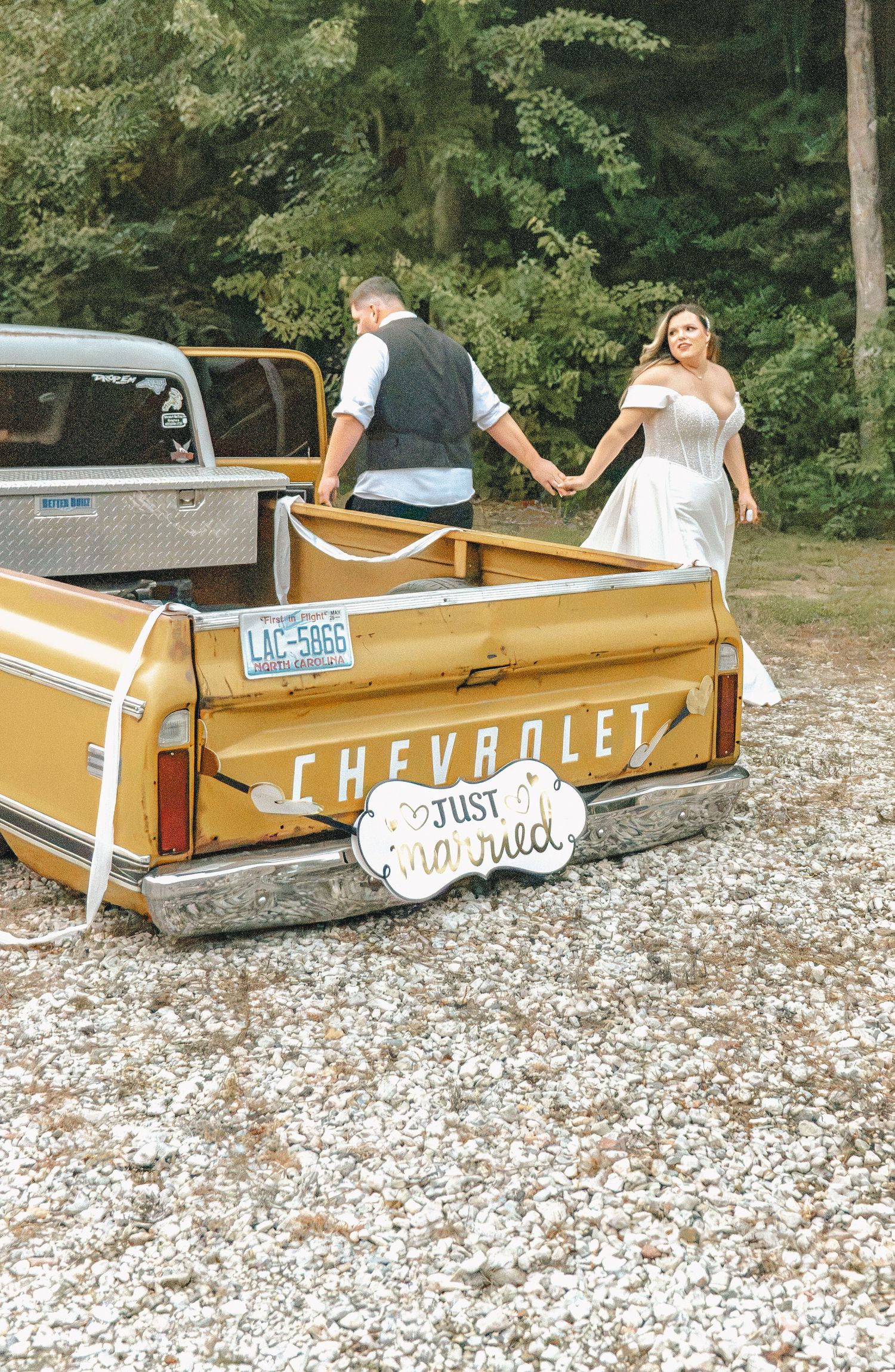 Newlywed couple holding hands in vintage yellow Chevrolet pickup truck bed with Just Married sign at rustic outdoor wedding.