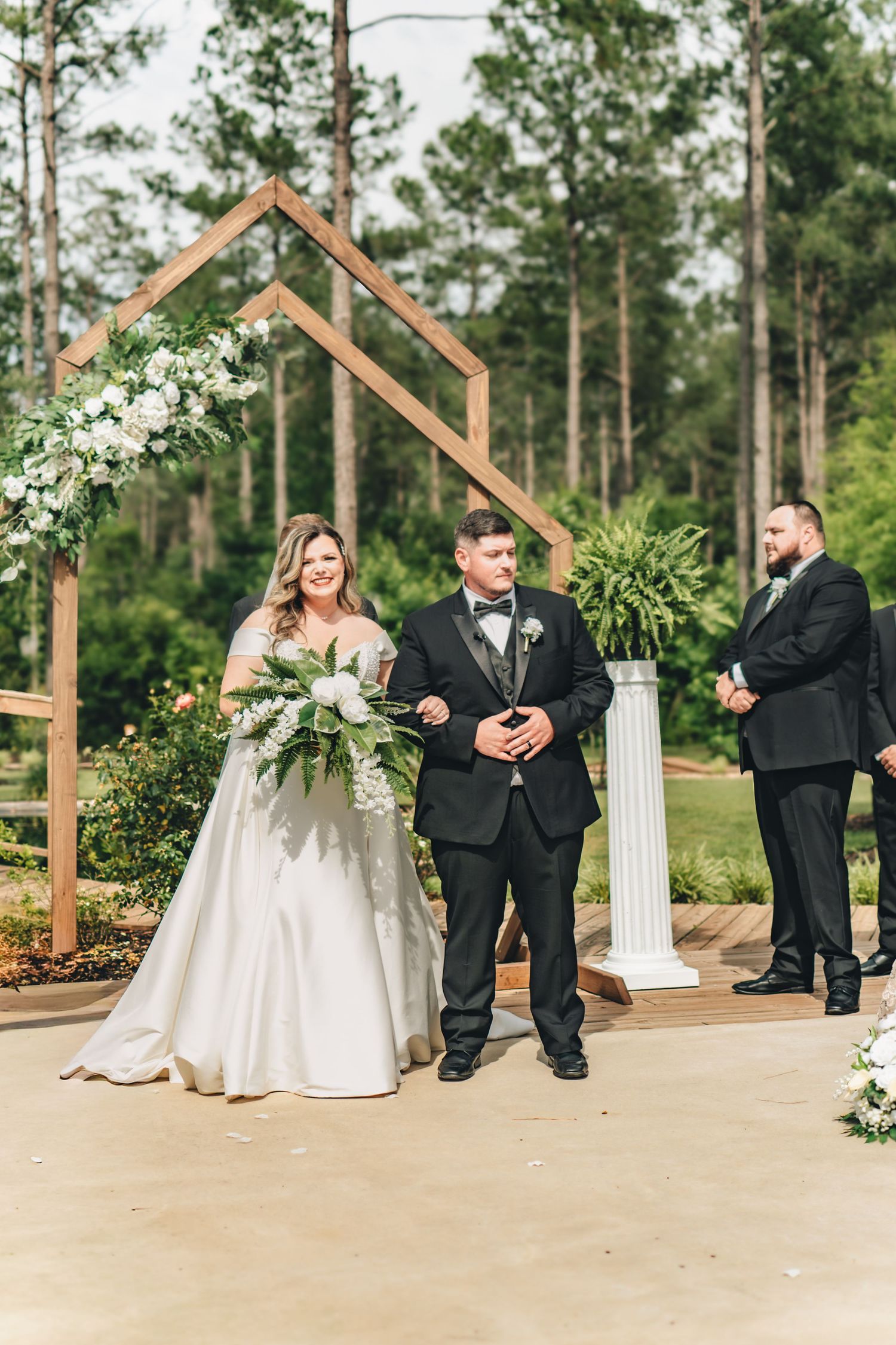 Bride and groom walk down the aisle at an outdoor wedding ceremony with a geometric wooden arch backdrop.