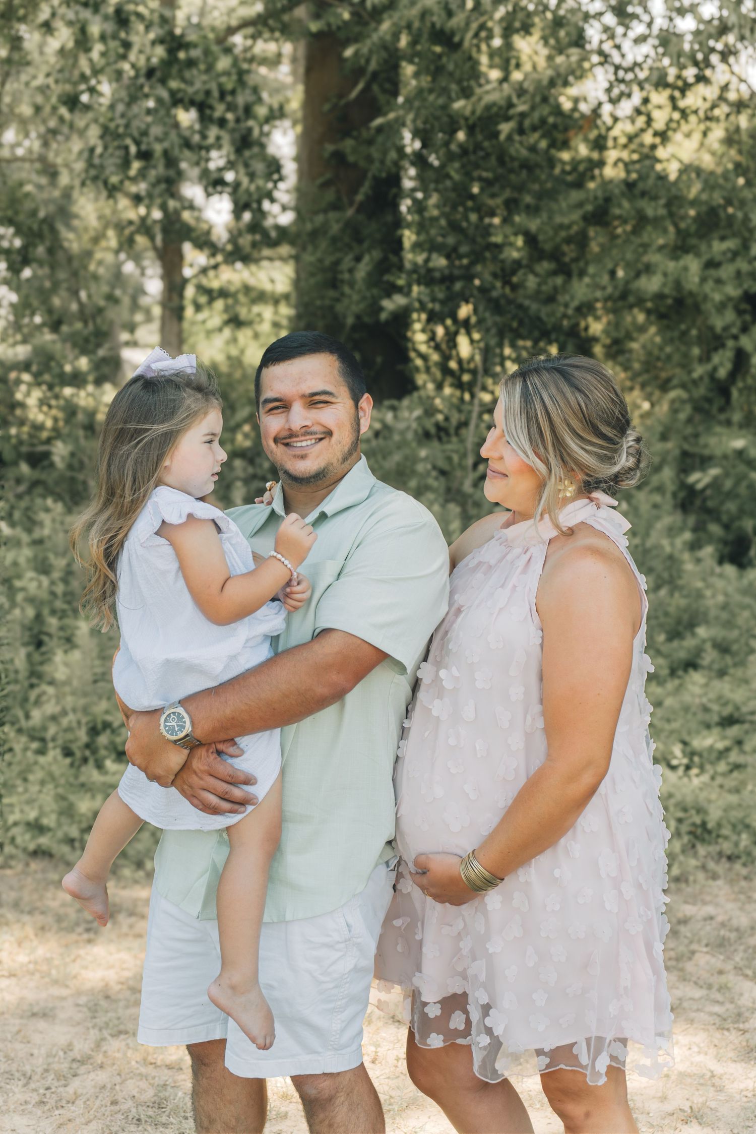 A smiling family with a man holding a toddler girl and a pregnant woman posing outdoors among green trees.