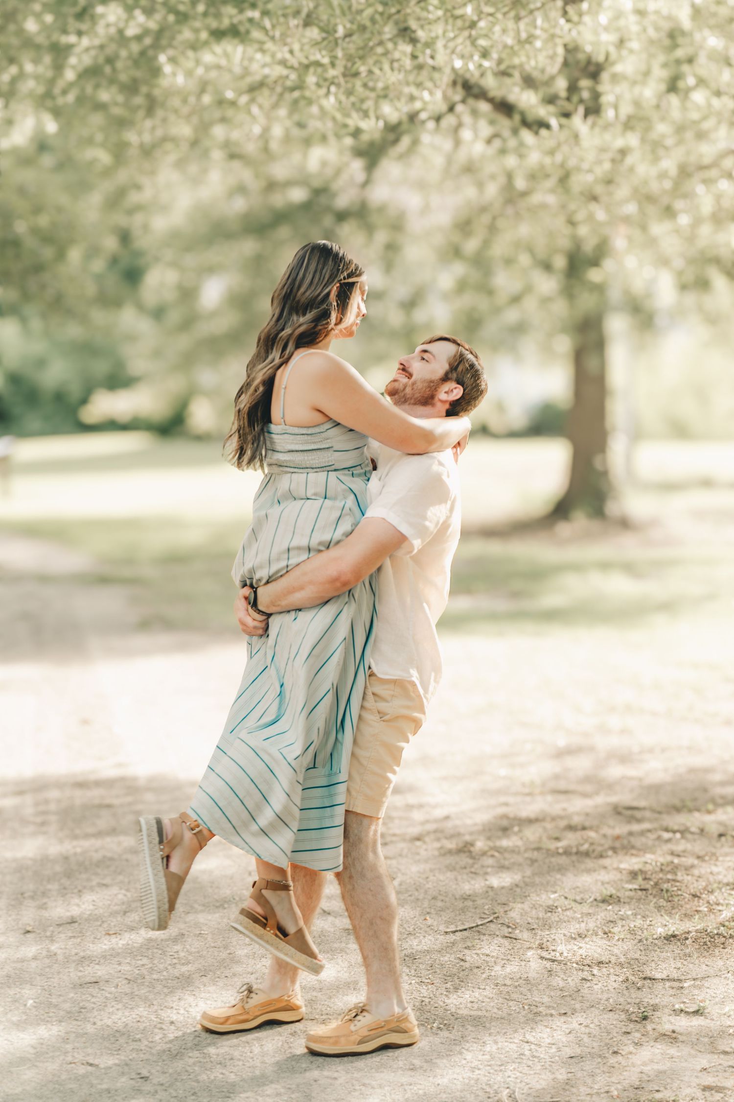 Couple embracing and laughing in a sunny park, woman in striped jumpsuit lifted by man in white shirt.