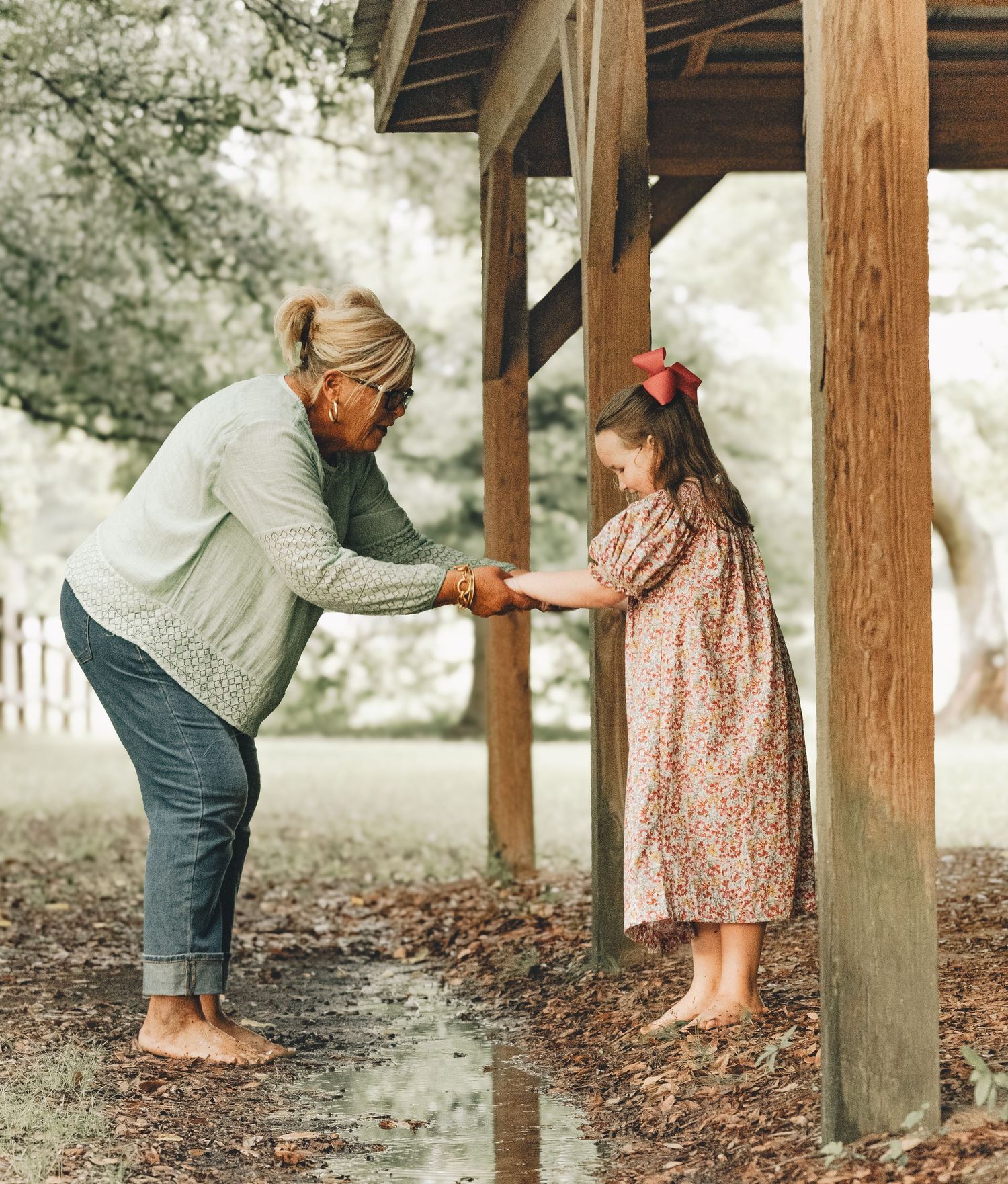 Woman bending down holding hands with a young girl in floral dress under a wooden pavilion outdoors.