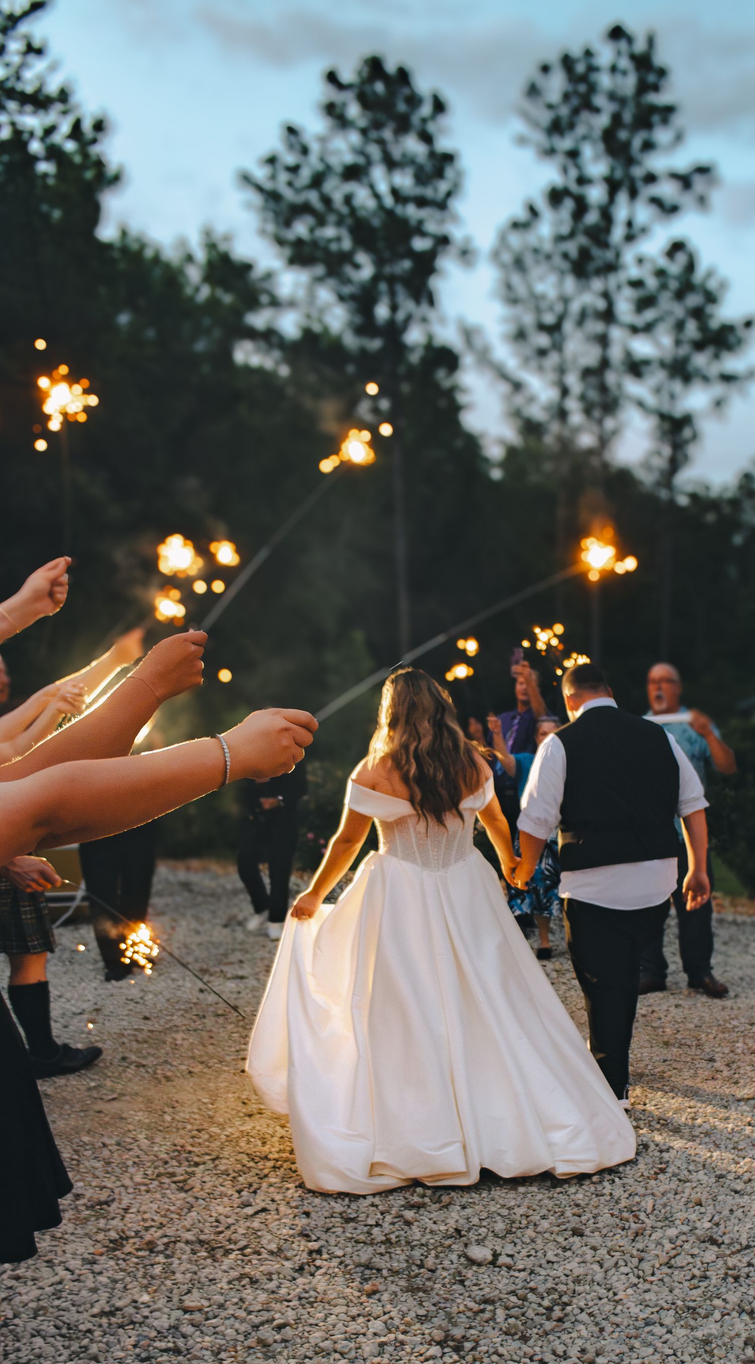 Bride and groom walk through sparkler send-off at outdoor evening wedding reception surrounded by guests.