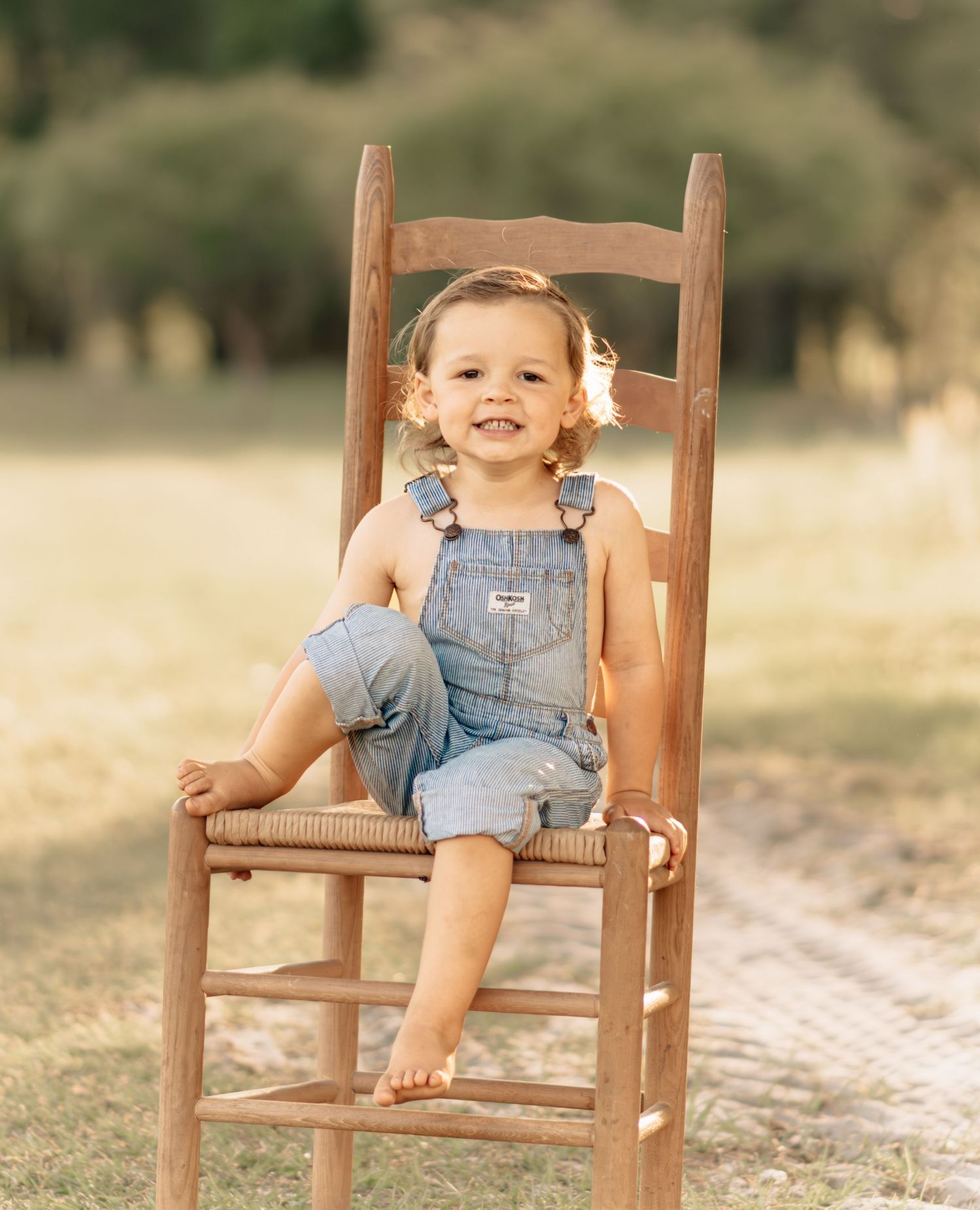Adorable toddler in denim overalls sitting on a rustic wooden chair smiling in an outdoor field setting.