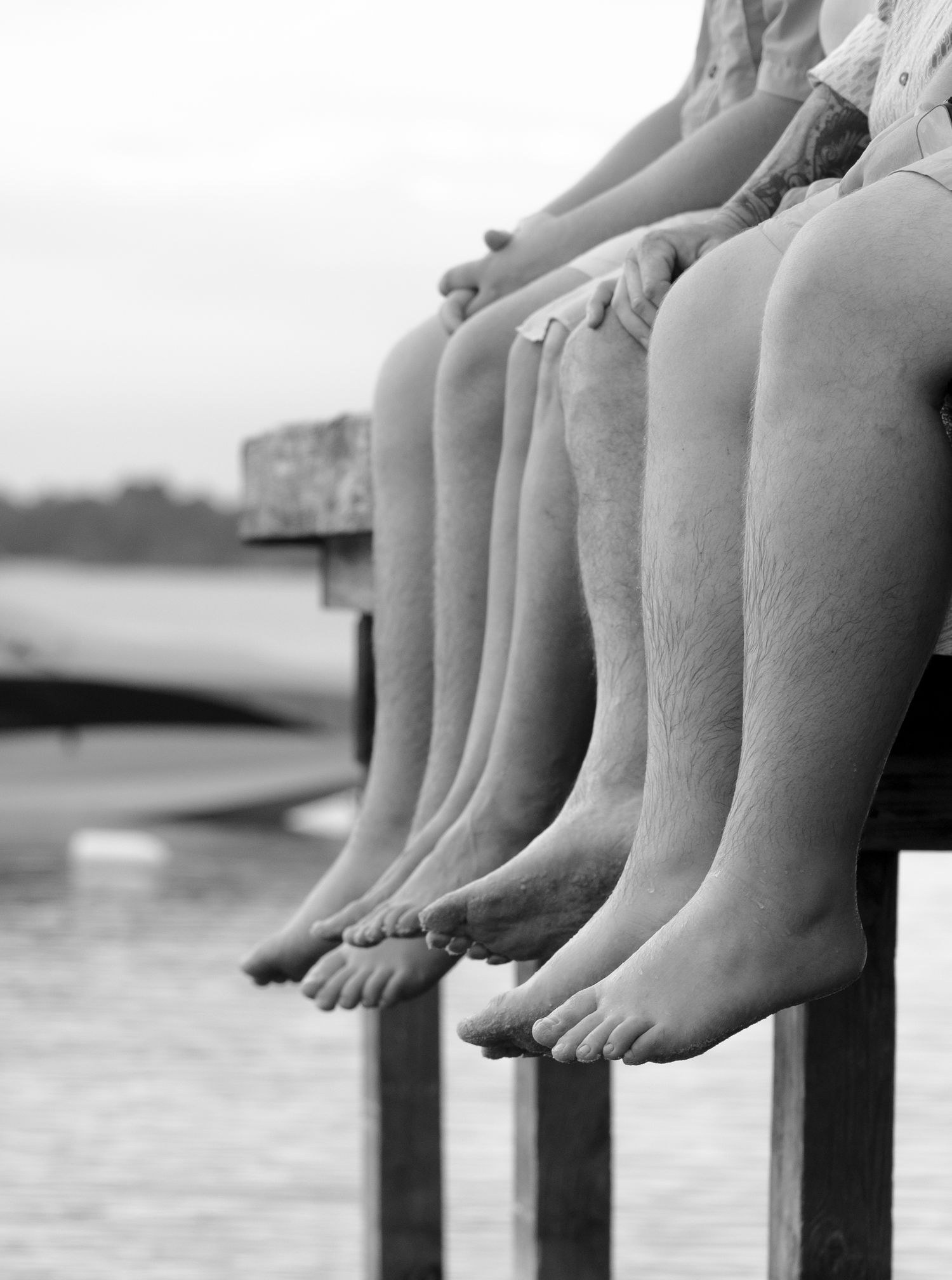 Black and white photo of a row of bare legs dangling off a wooden dock over water.