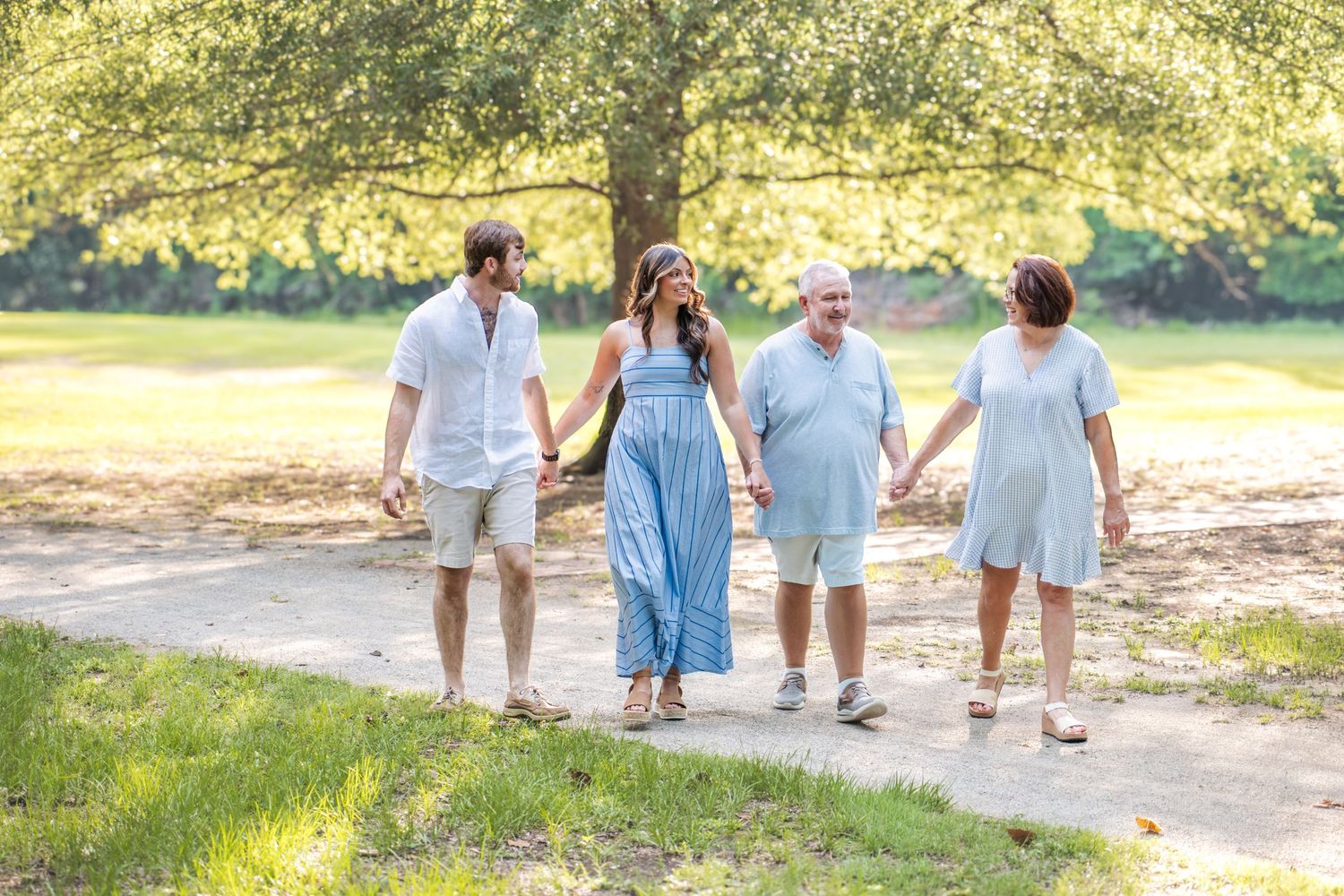 Four family members walk hand-in-hand on a sunny park path surrounded by lush green trees.