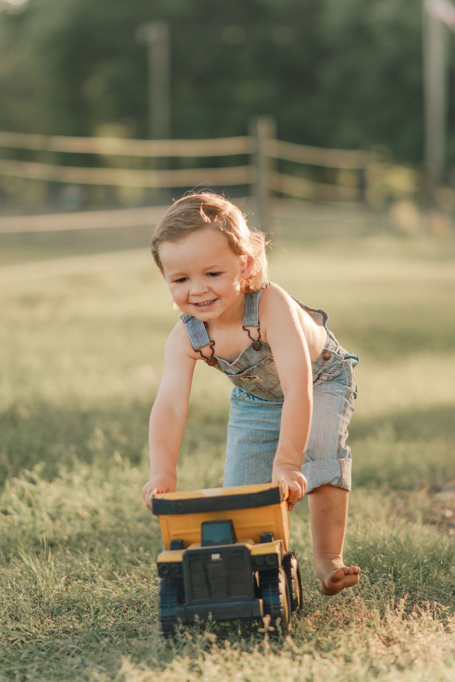 Joyful toddler in denim overalls pushing a yellow toy truck across a green grassy field at golden hour.