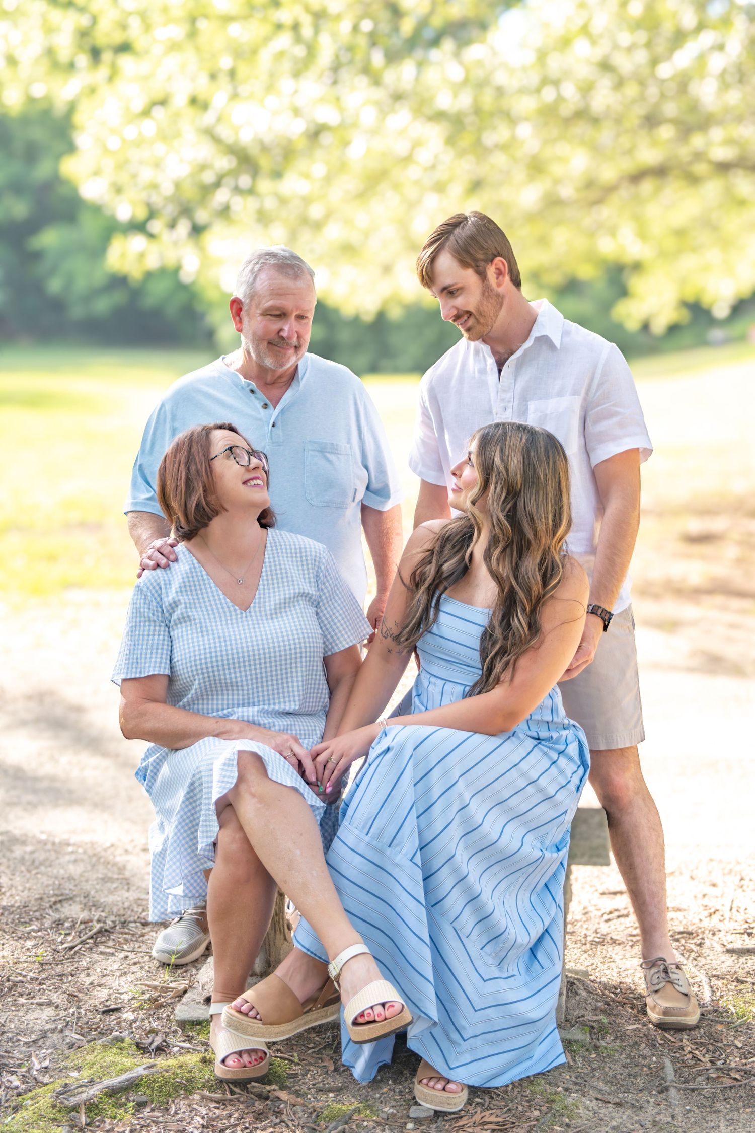 Two couples pose outdoors in coordinating blue and white outfits, smiling at each other in a sunlit park setting.