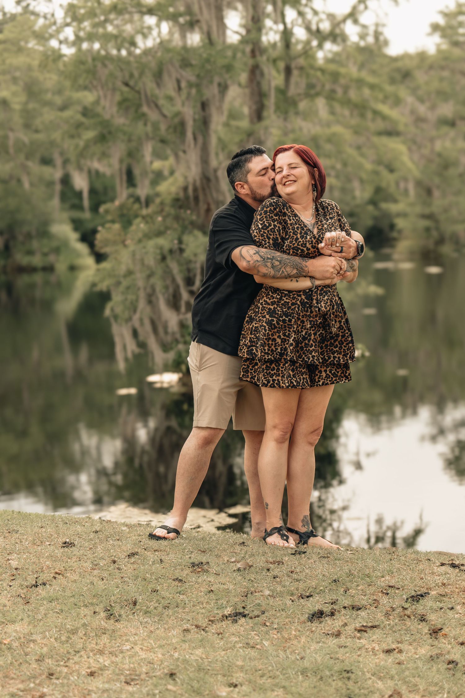 A couple embraces near a serene lake surrounded by cypress trees, the man kissing the smiling woman in a leopard print dress.
