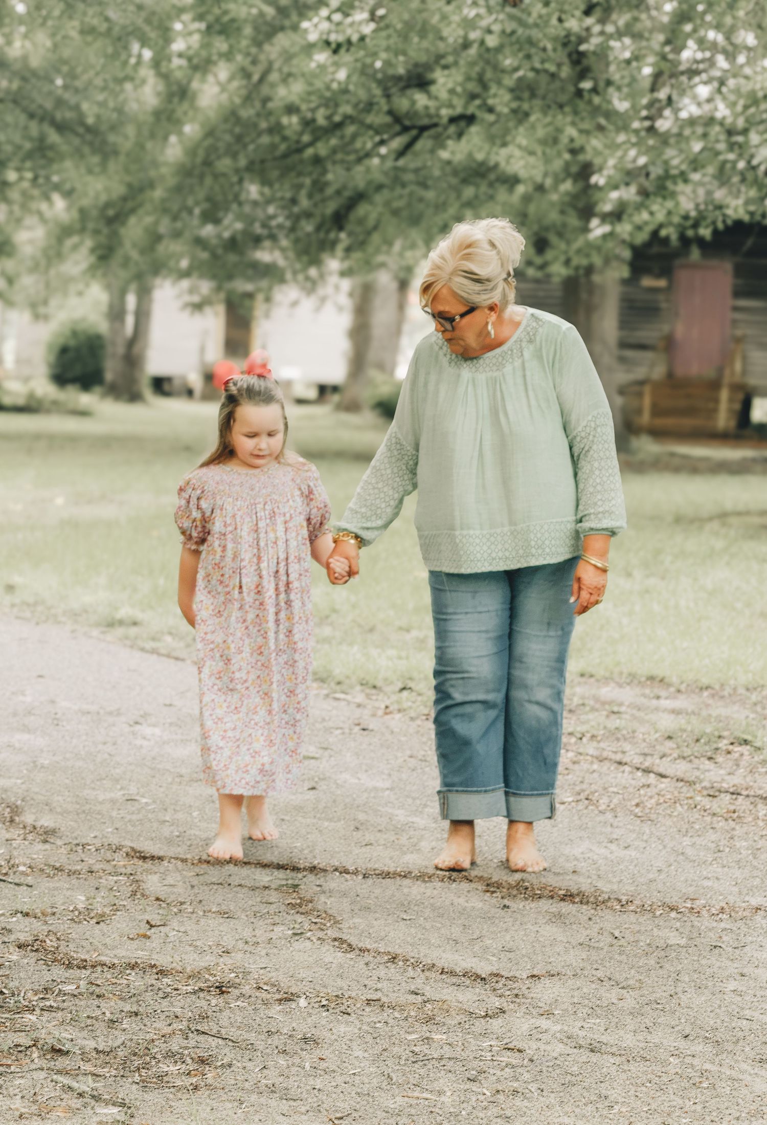An older woman in a mint blouse and jeans holds hands with a young girl in a floral dress while walking on a dirt path.