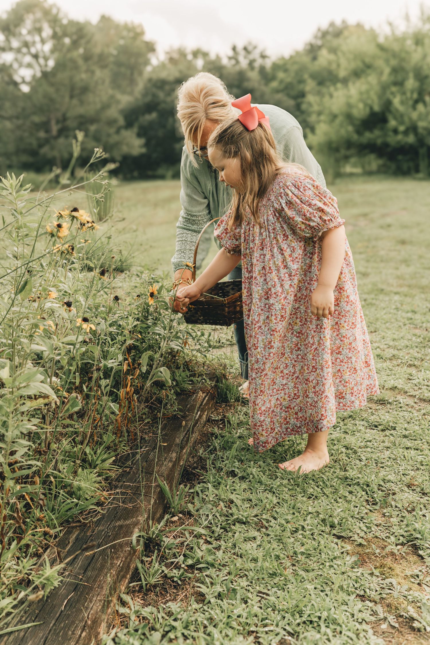 A woman and young girl in a floral dress pick flowers together in a lush garden, placing them in a wicker basket.