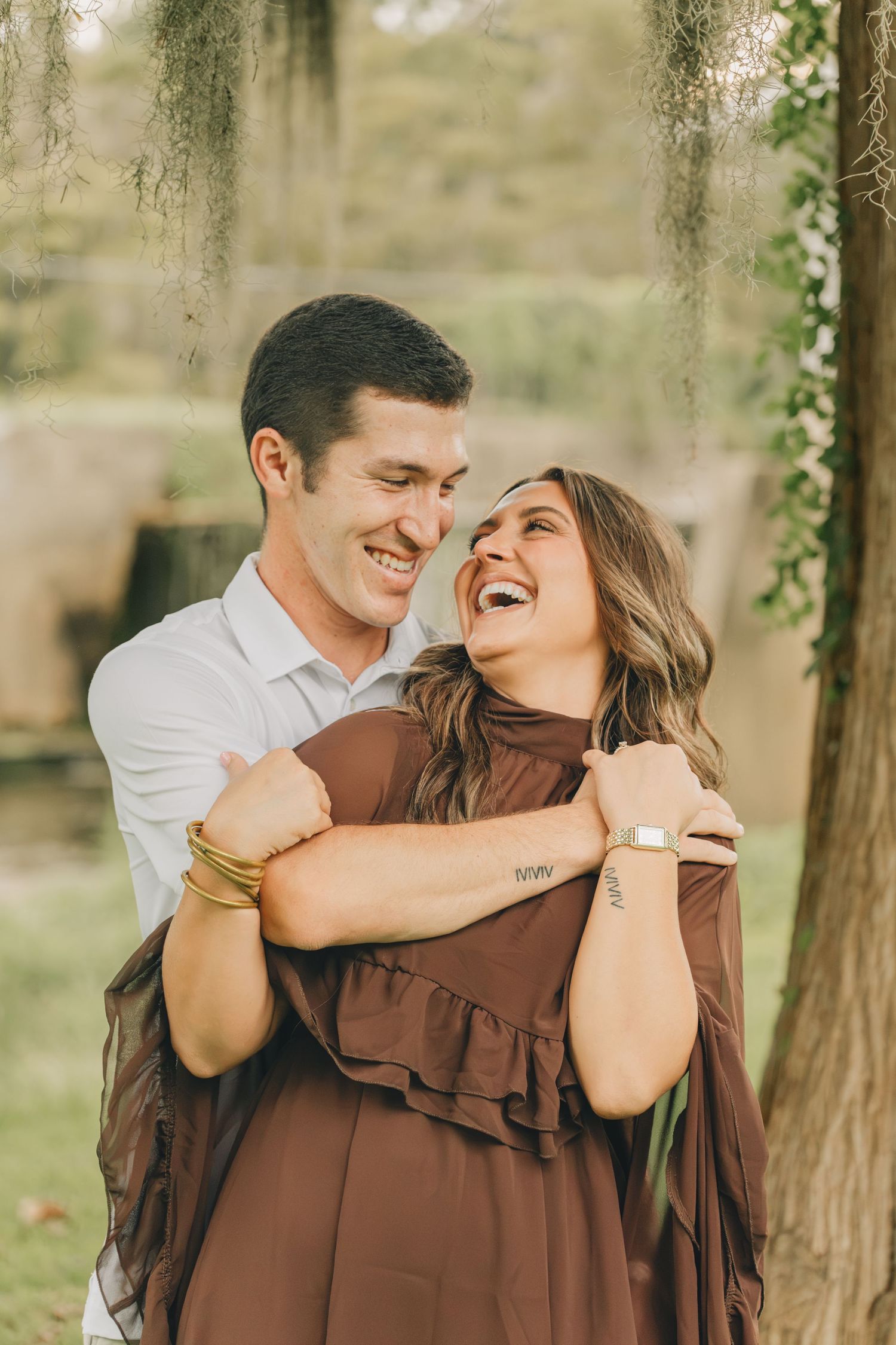 A laughing couple embraces outdoors, woman in brown dress, man in white shirt, surrounded by lush greenery and hanging moss.