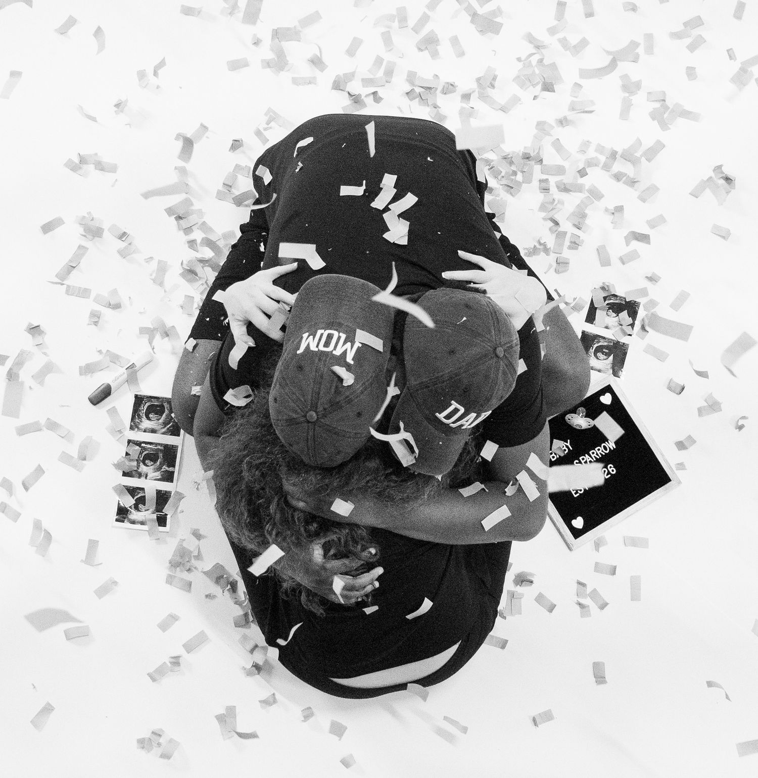 Aerial black and white photo of couple embracing surrounded by confetti and ultrasound photos on white floor.