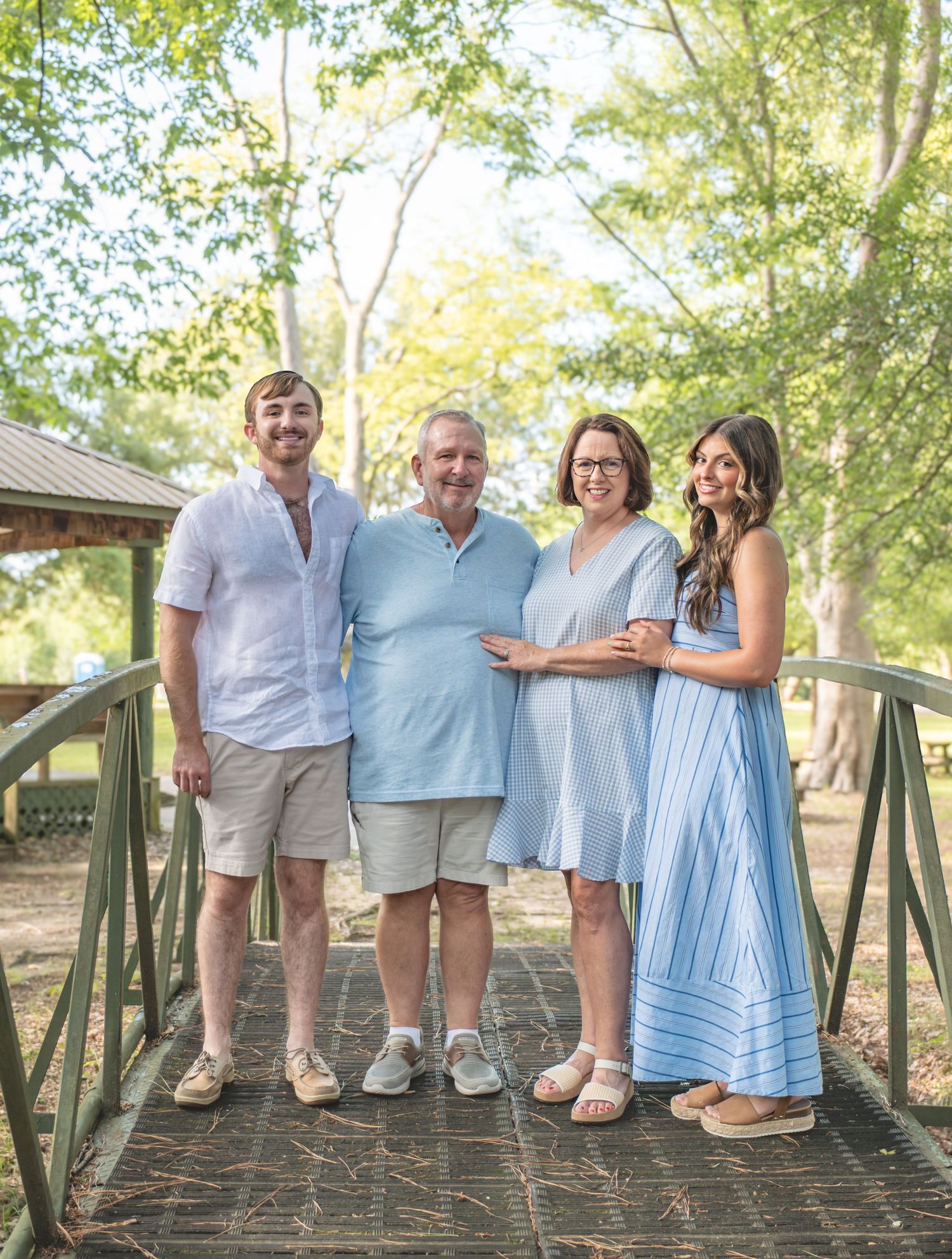 Happy family of four smiling together on a wooden bridge surrounded by lush green trees in coordinated blue outfits.