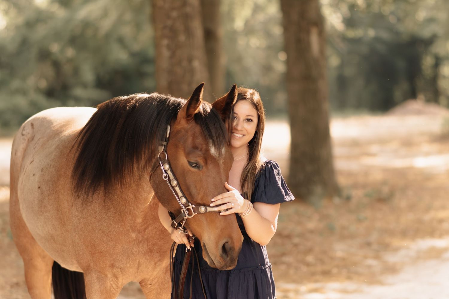 Young brunette woman posing with brown horse on shaded dirt path outdoors.