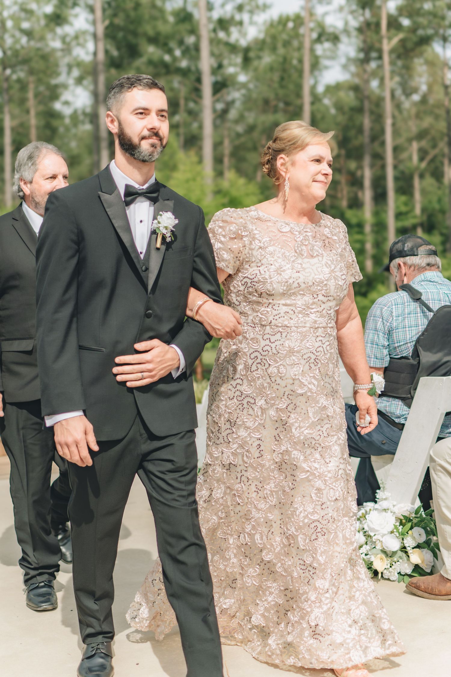 Groom escorts elegantly dressed mother down wedding aisle in lace gown during outdoor ceremony.
