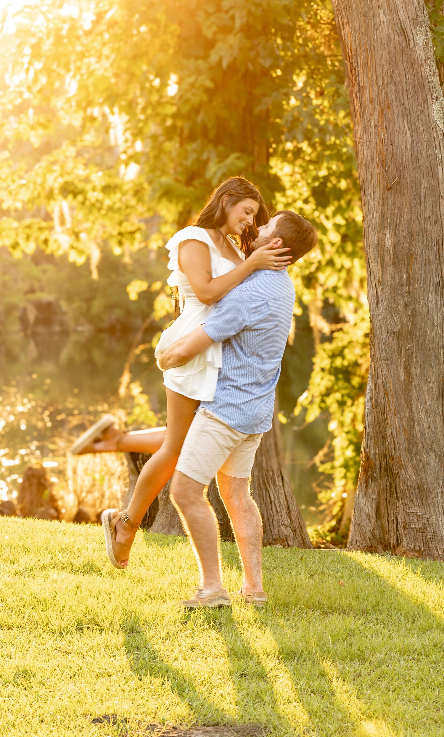 A couple embraces and lifts in golden sunset light beside a tree in a lush green park.