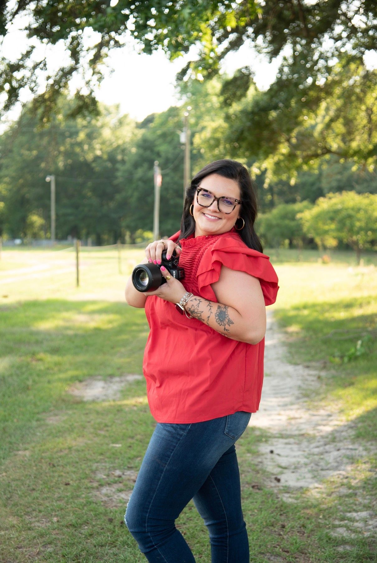 A smiling woman with glasses holds a DSLR camera, wearing a red top and jeans, standing outdoors in a green park.