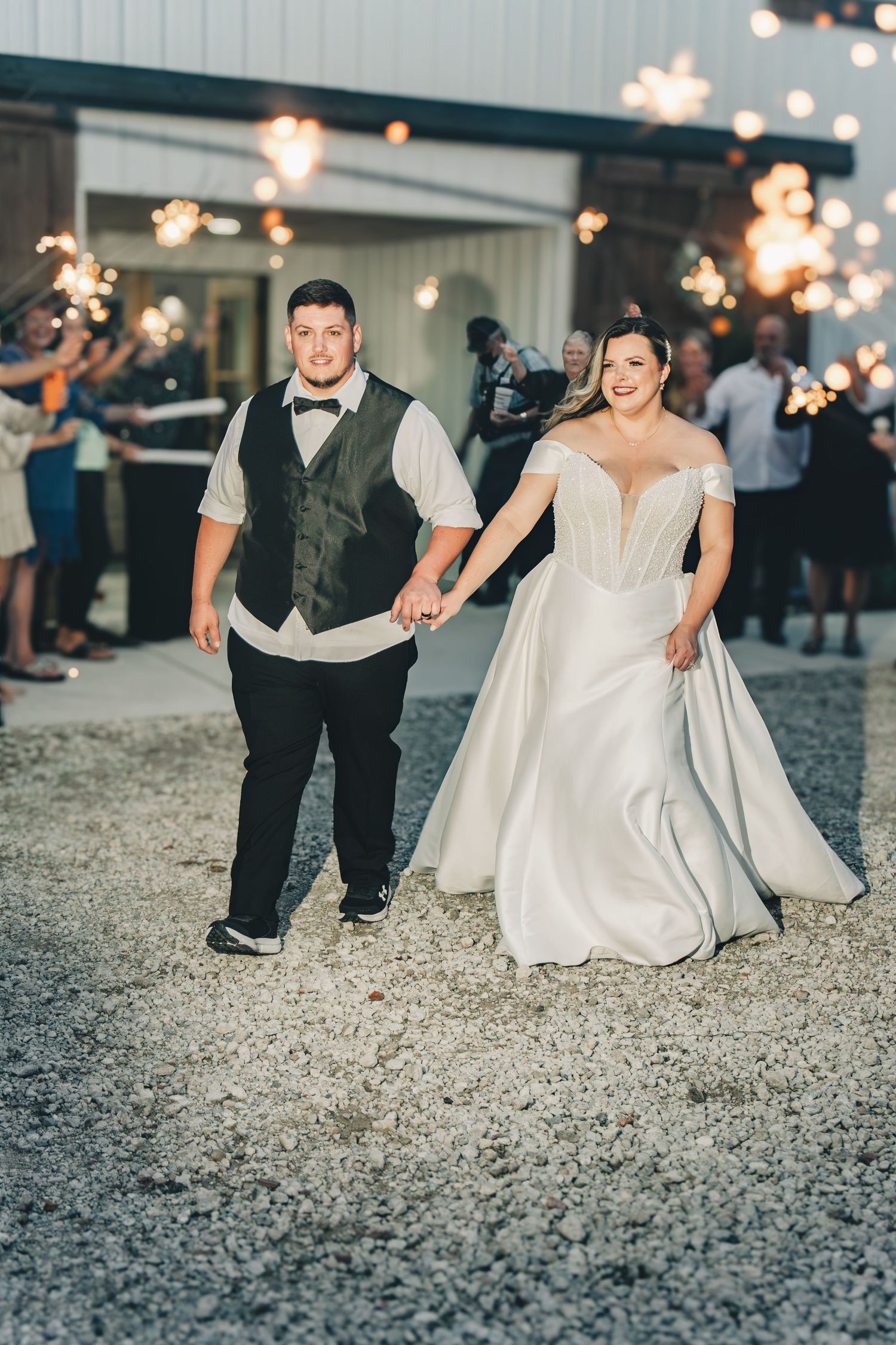 Bride in off-shoulder gown and groom in vest hold hands during sparkler exit at outdoor barn wedding reception.