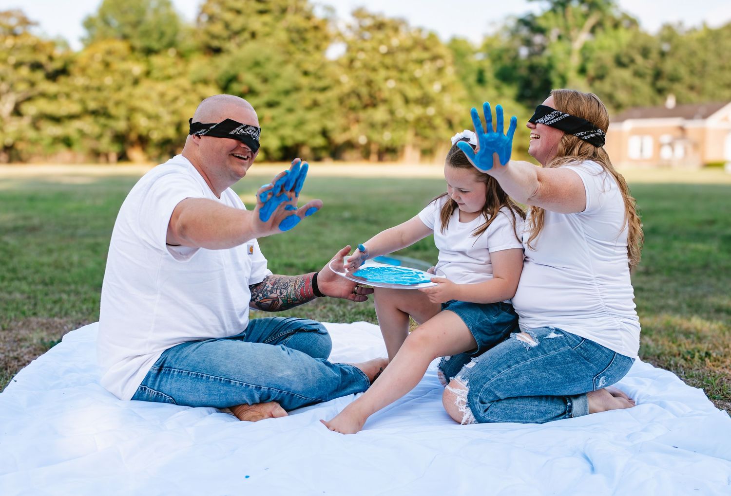 Family of three with blue paint on hands sitting on white blanket outdoors, smiling and playing together.