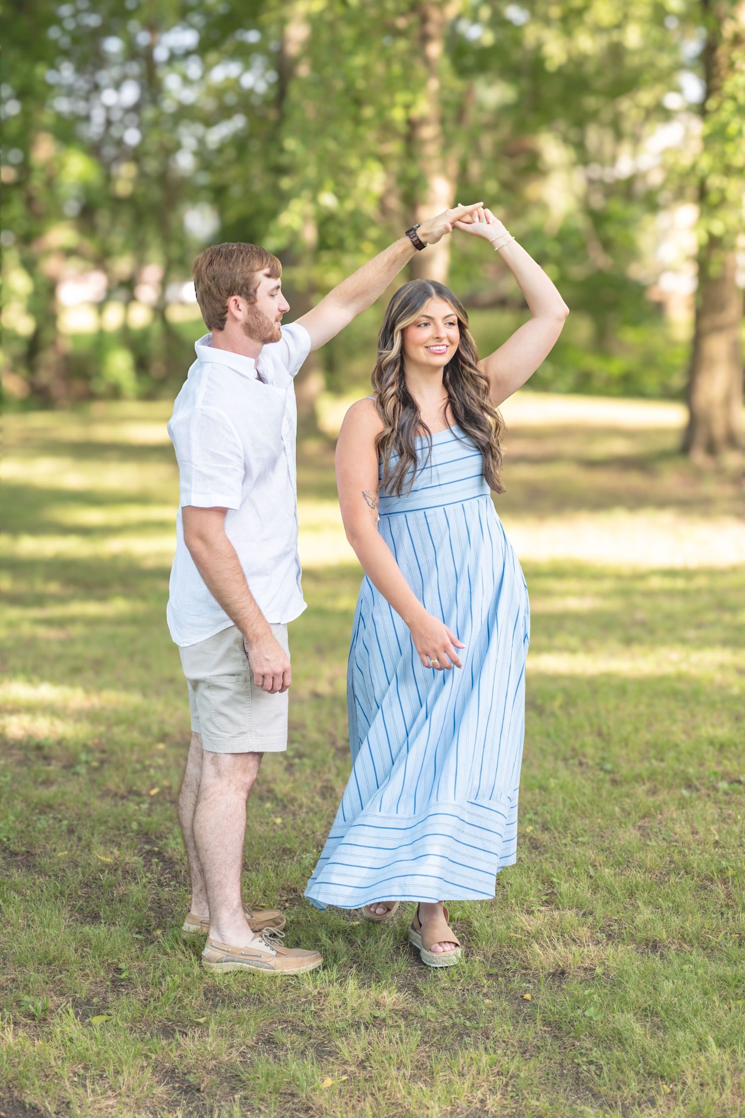Man spinning woman in blue striped maxi dress while dancing together in a sunny green park.
