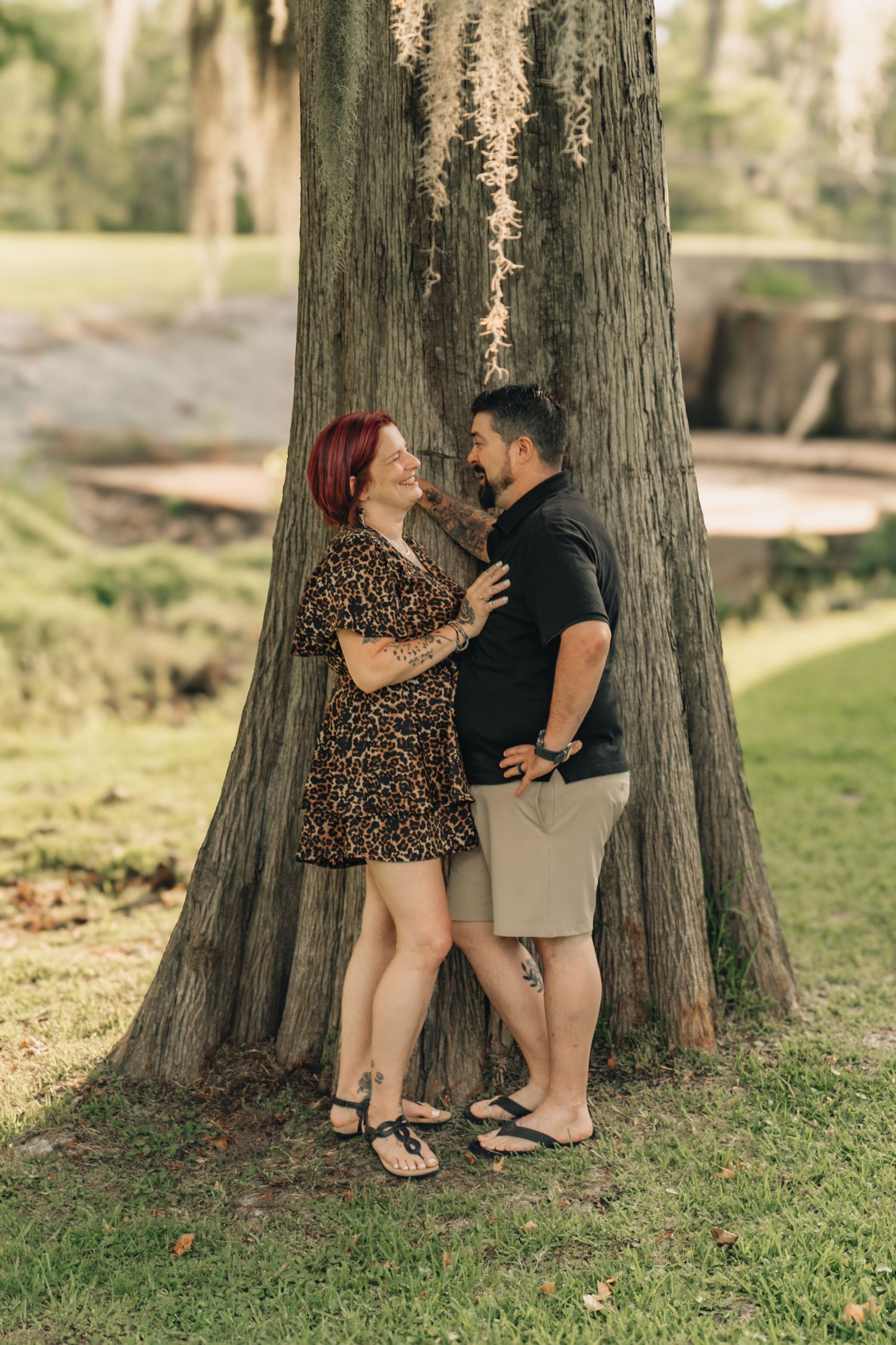 Couple sharing a romantic moment leaning against a large tree in a lush green park setting.