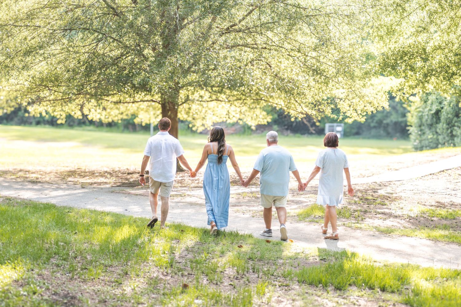 Family of four holding hands walking away on a sunny path surrounded by lush green trees.