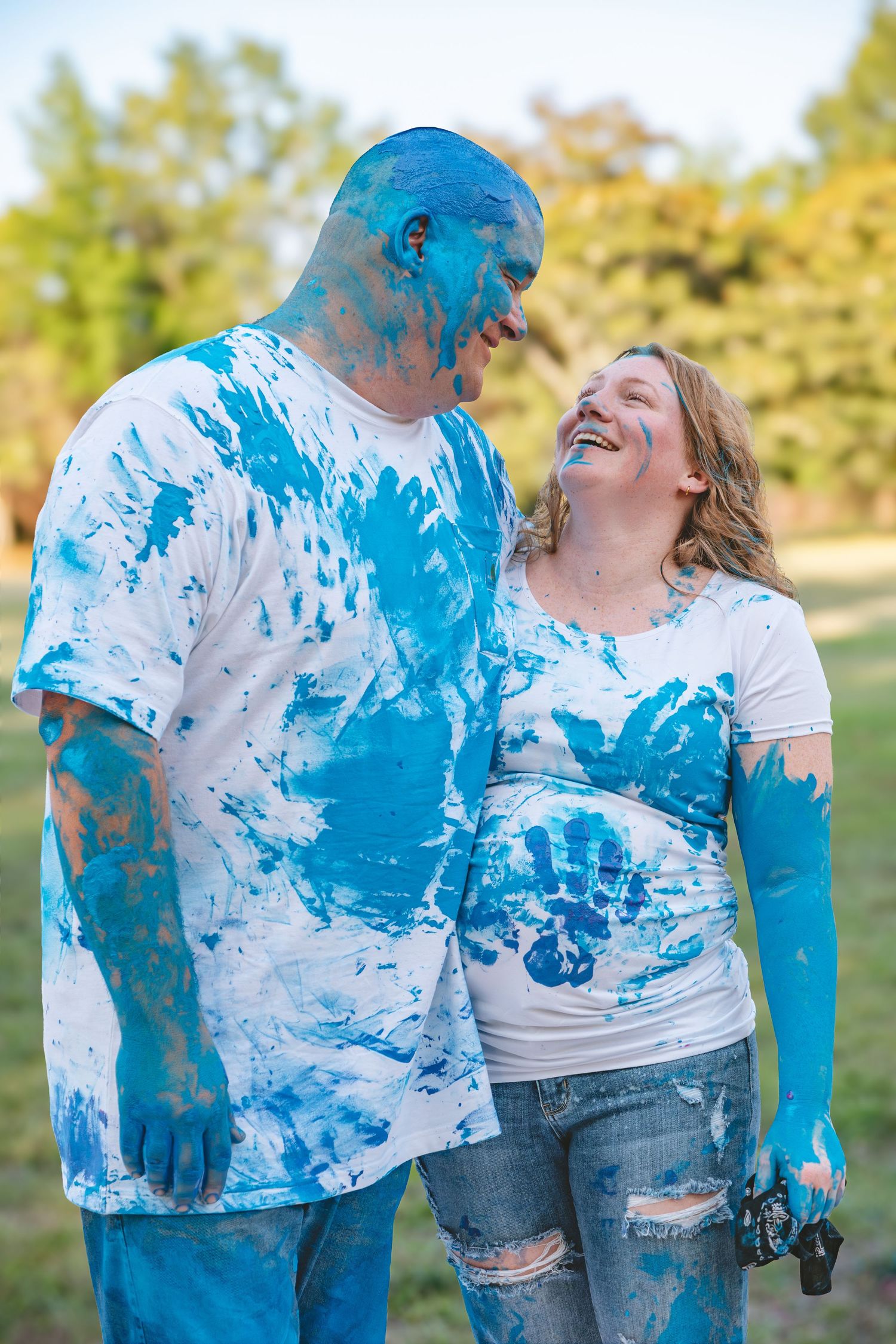 Couple covered in blue paint smiling at each other outdoors, celebrating a gender reveal with blue handprints on white shirts.