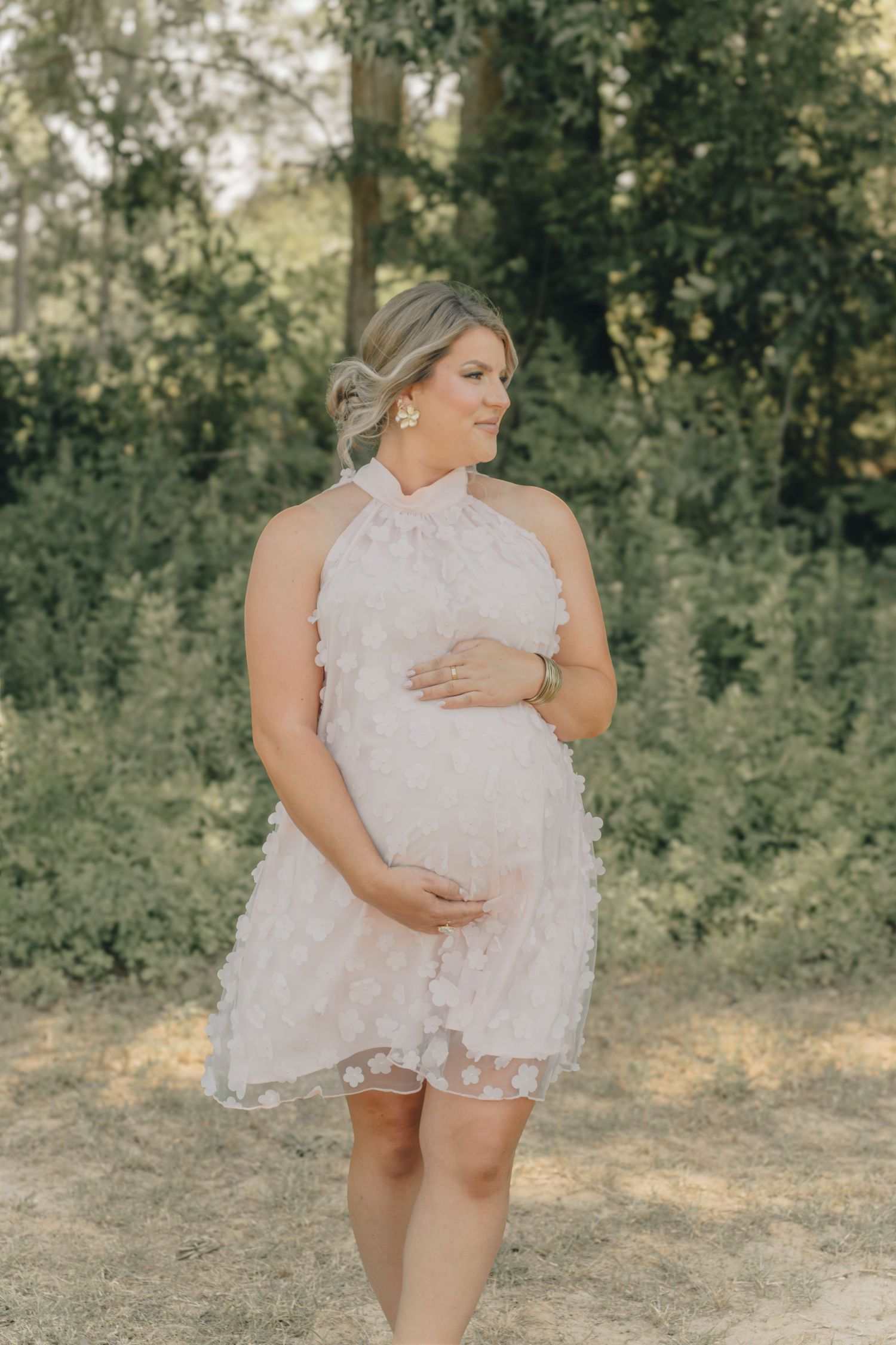 Pregnant woman in a blush floral halter dress cradling her baby bump outdoors in a lush green garden setting.