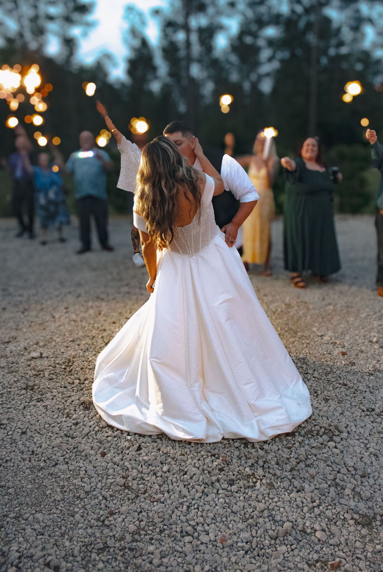 Newlywed couple sharing a kiss in wedding attire surrounded by guests holding sparklers at dusk.
