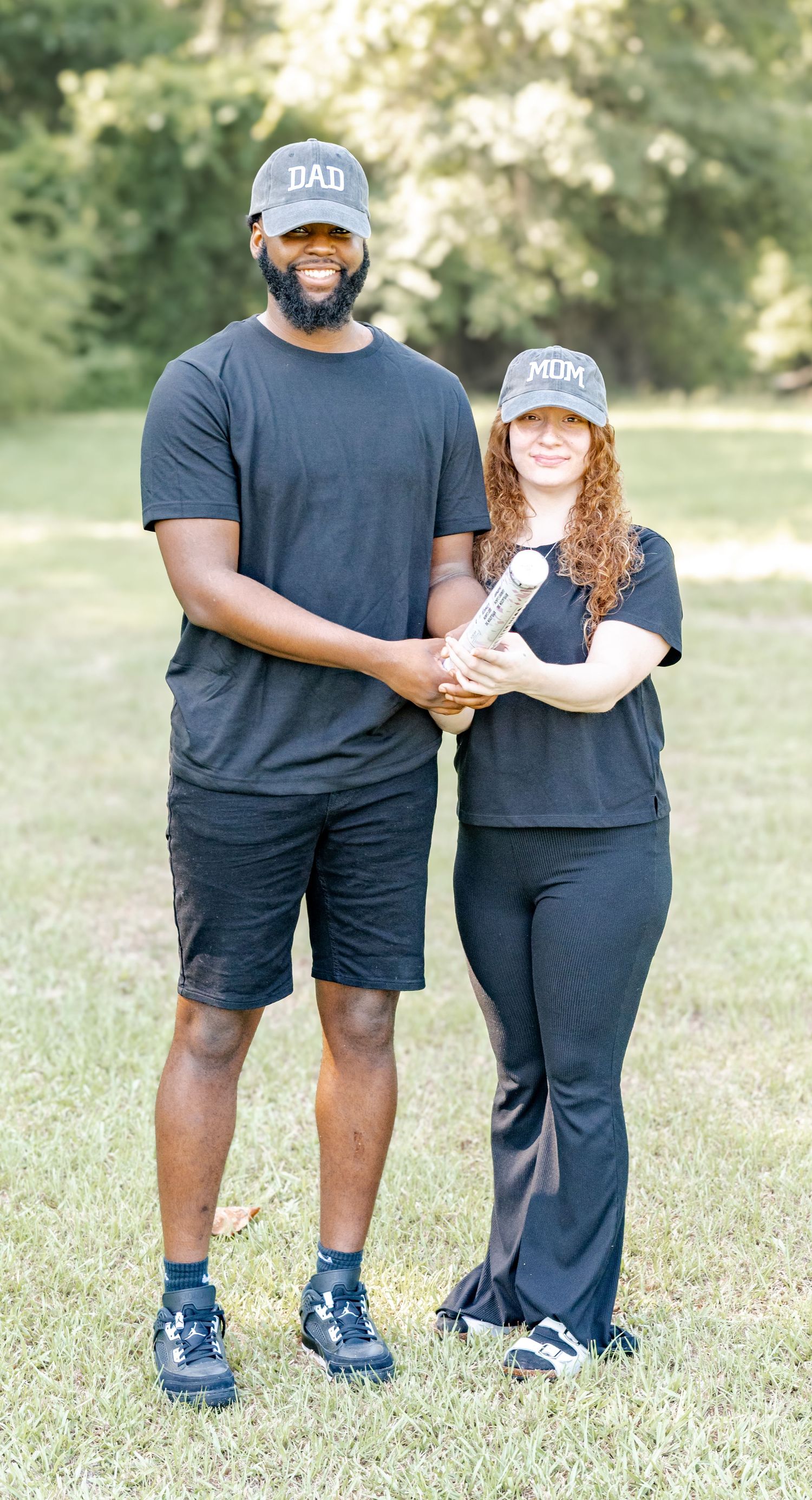Smiling couple wearing matching DAD and MOM caps holding a cylinder together in a sunny outdoor park.