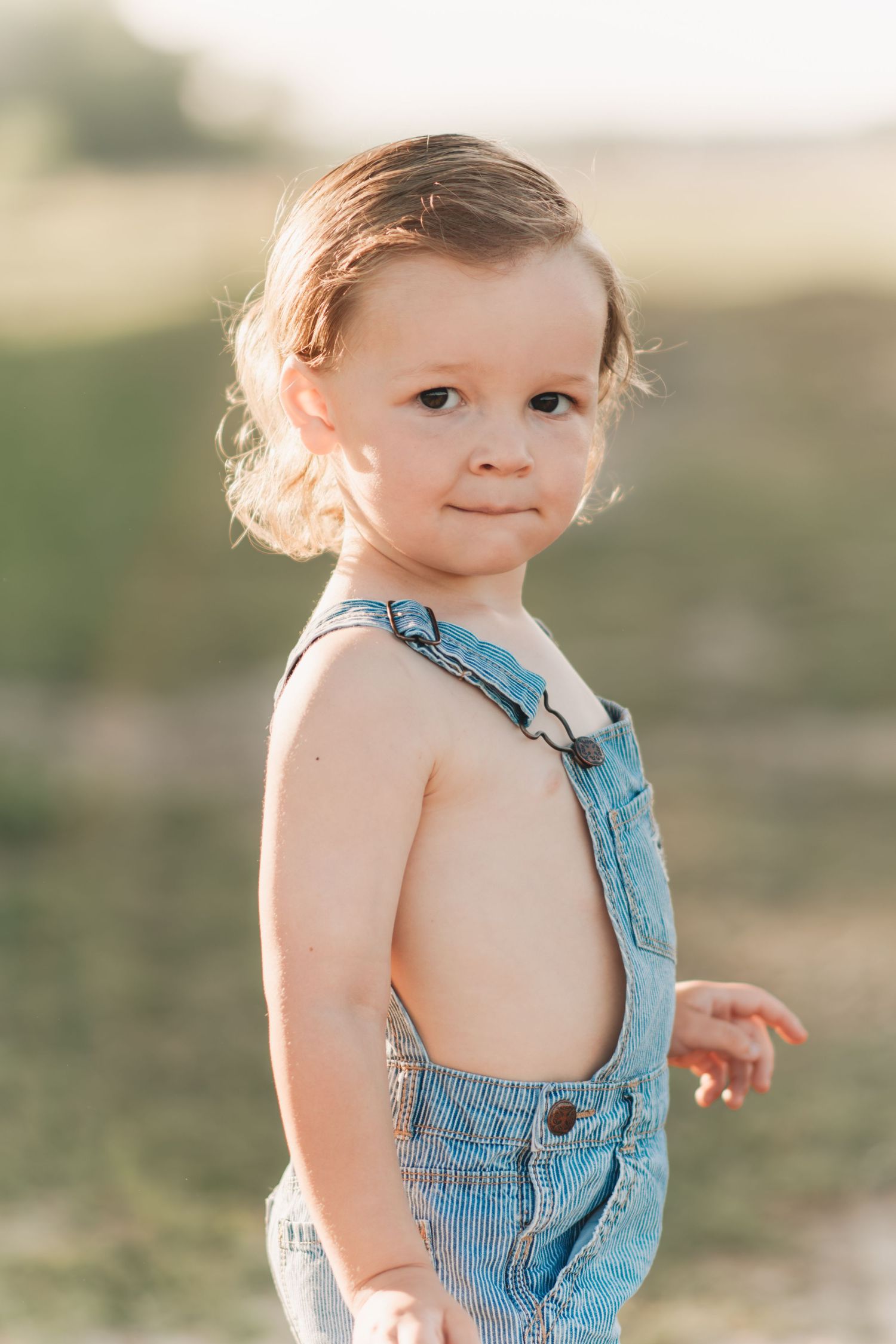 Toddler in denim overalls smiling outdoors in golden sunlight.