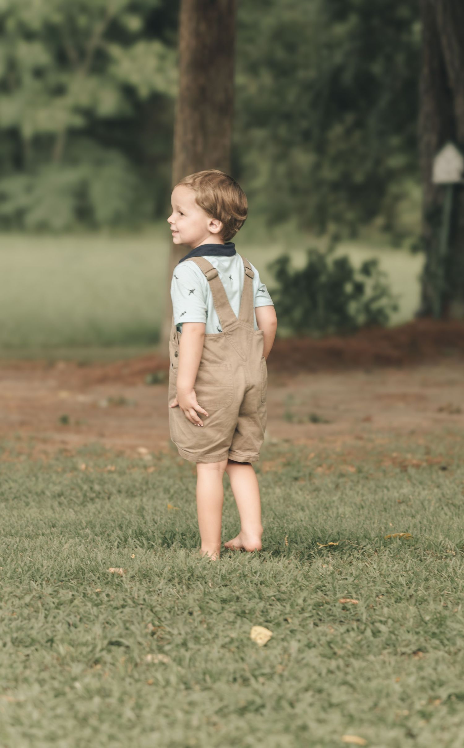 A toddler boy in tan overalls and light blue shirt stands barefoot on green grass outdoors.