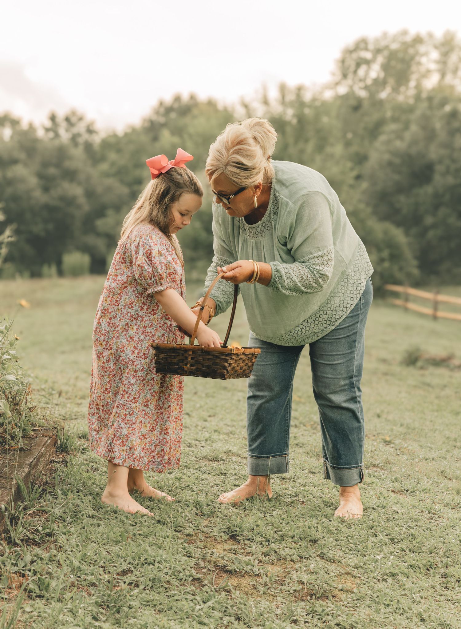 A grandmother and young girl in floral dress examine a wicker basket together in a lush green field.