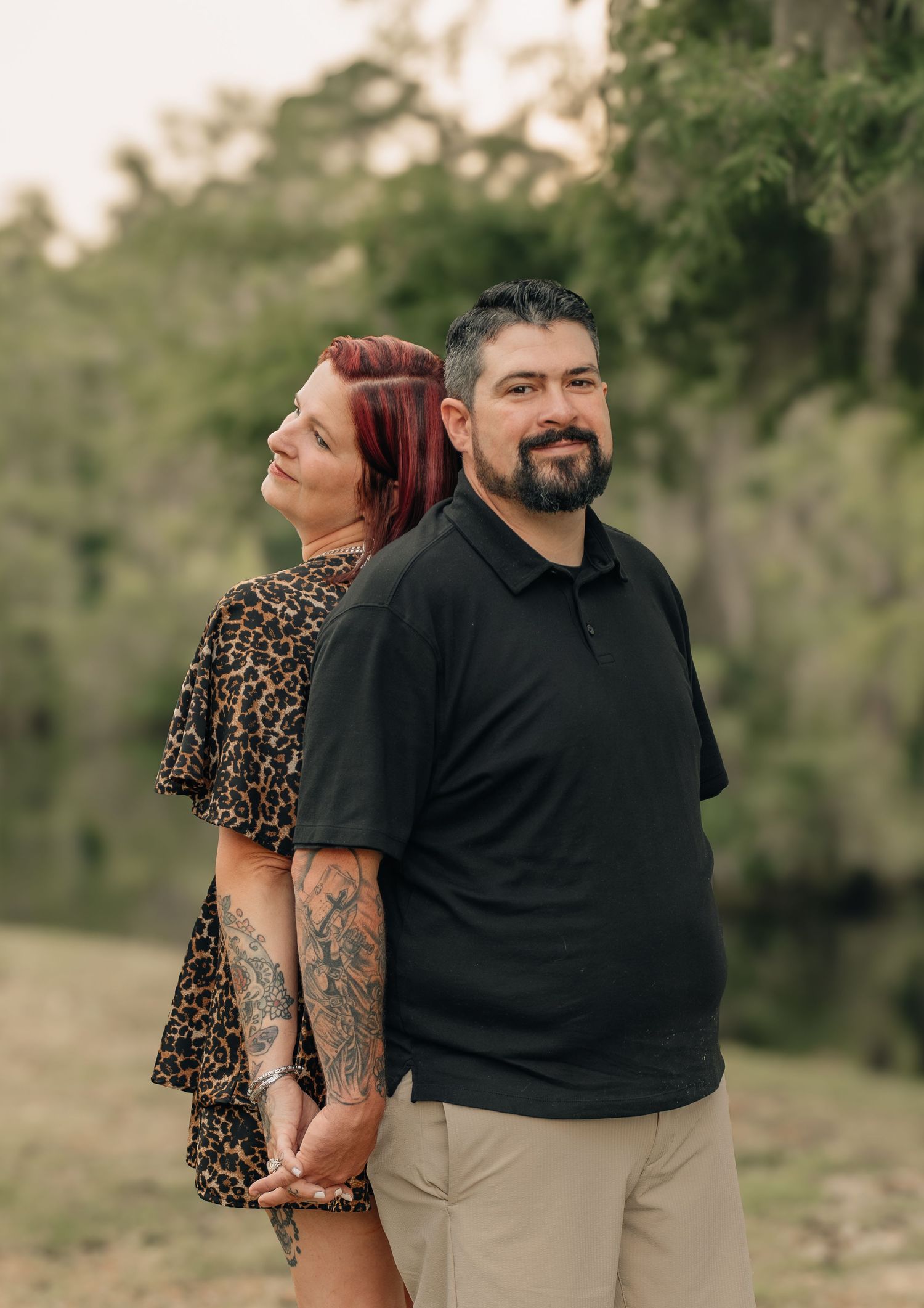Couple posing back-to-back outdoors near a river, woman with red hair and tattoos in leopard dress, man in black polo.