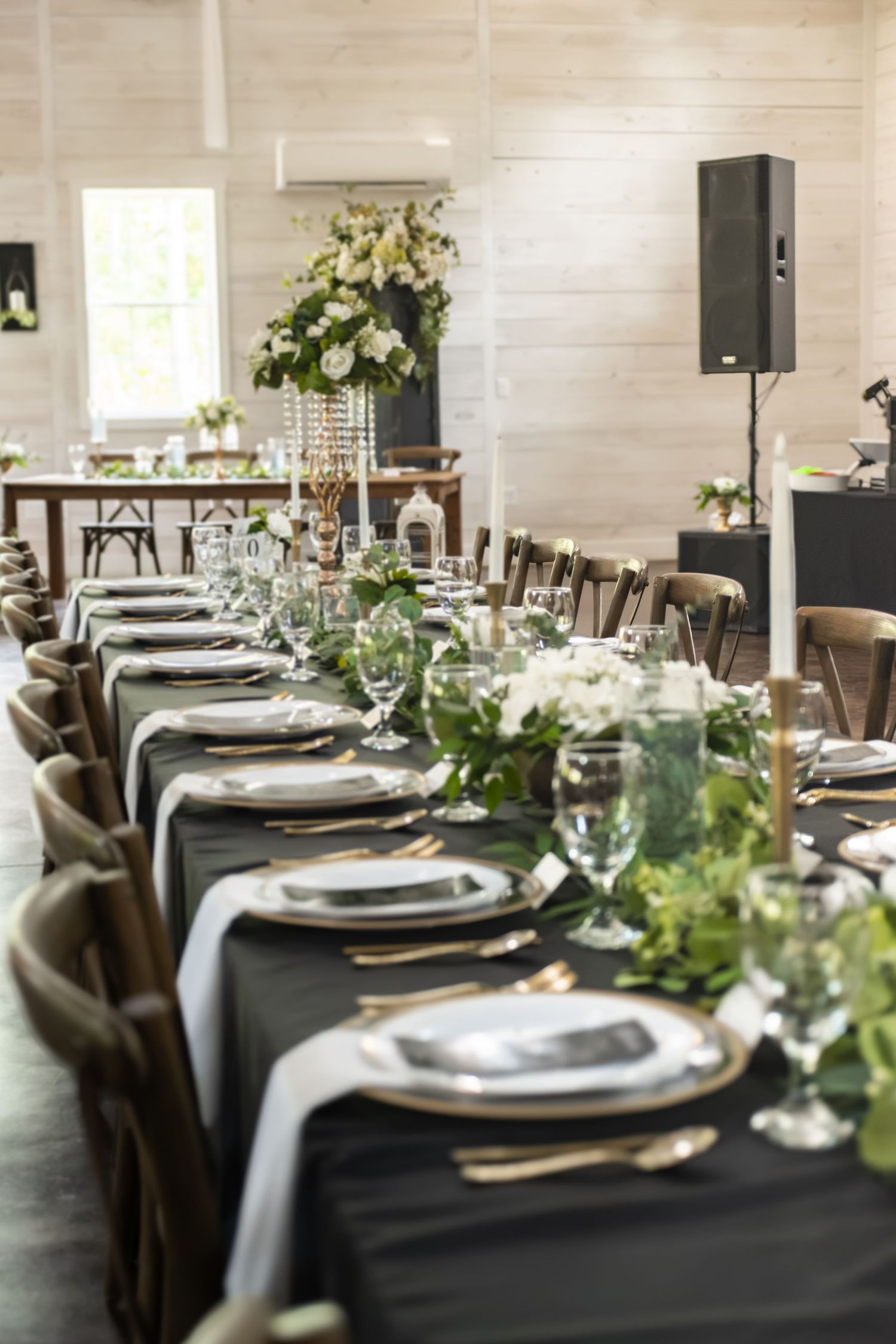 Elegant wedding reception table with dark linens, gold accents, greenery centerpieces, and tall floral arrangements in a white barn venue.