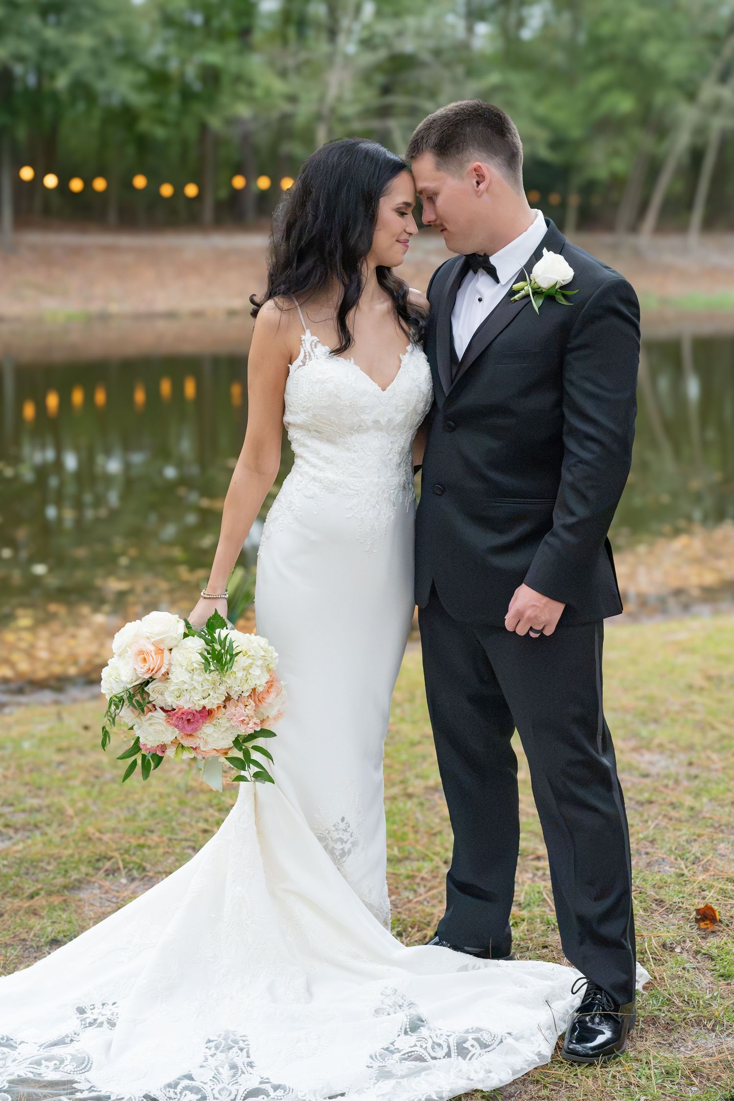 Bride in a lace wedding gown holding a pastel bouquet embraces a groom in a black tuxedo beside a pond with string lights.