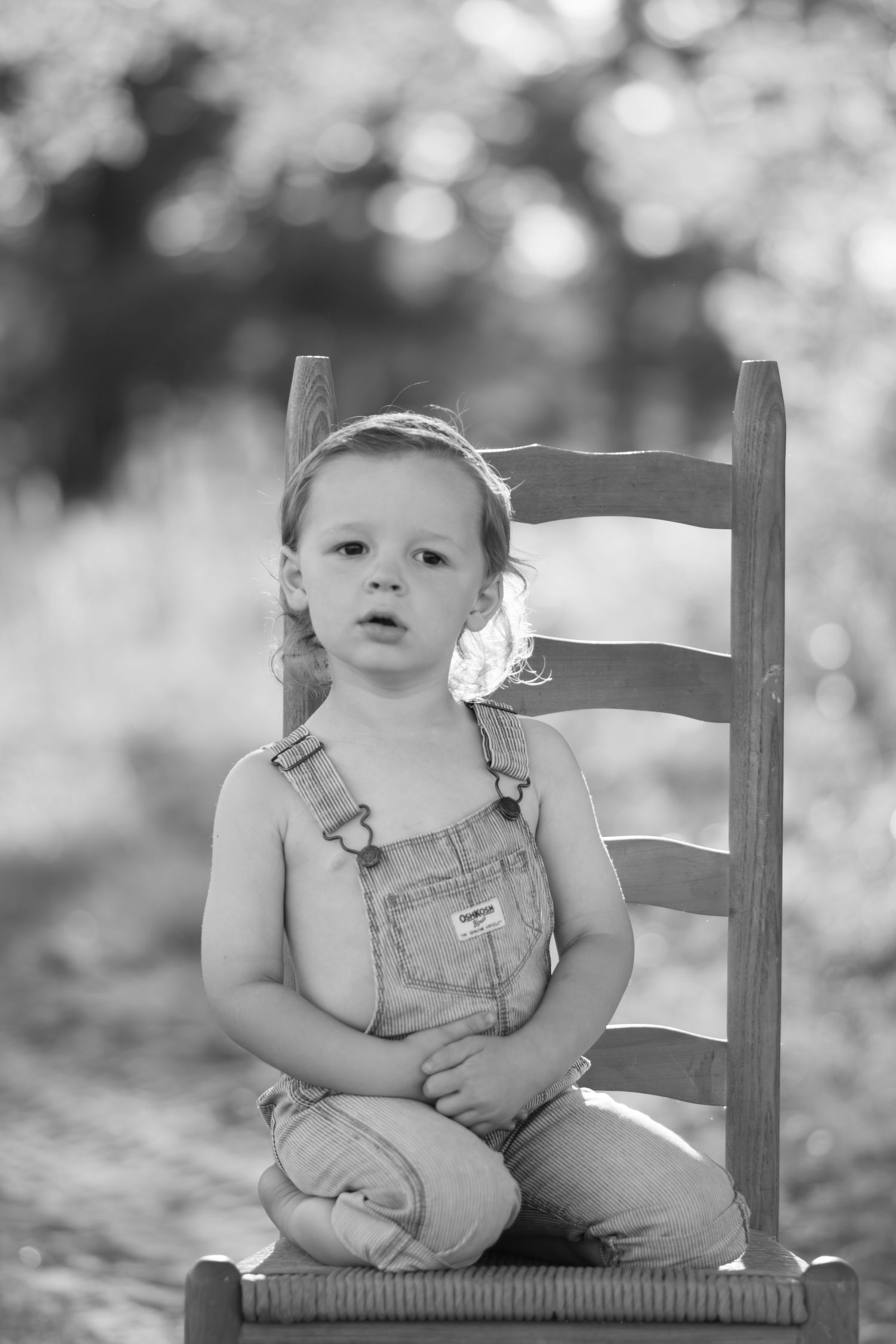 Black and white portrait of a young toddler in denim overalls sitting quietly on a wooden chair outdoors.