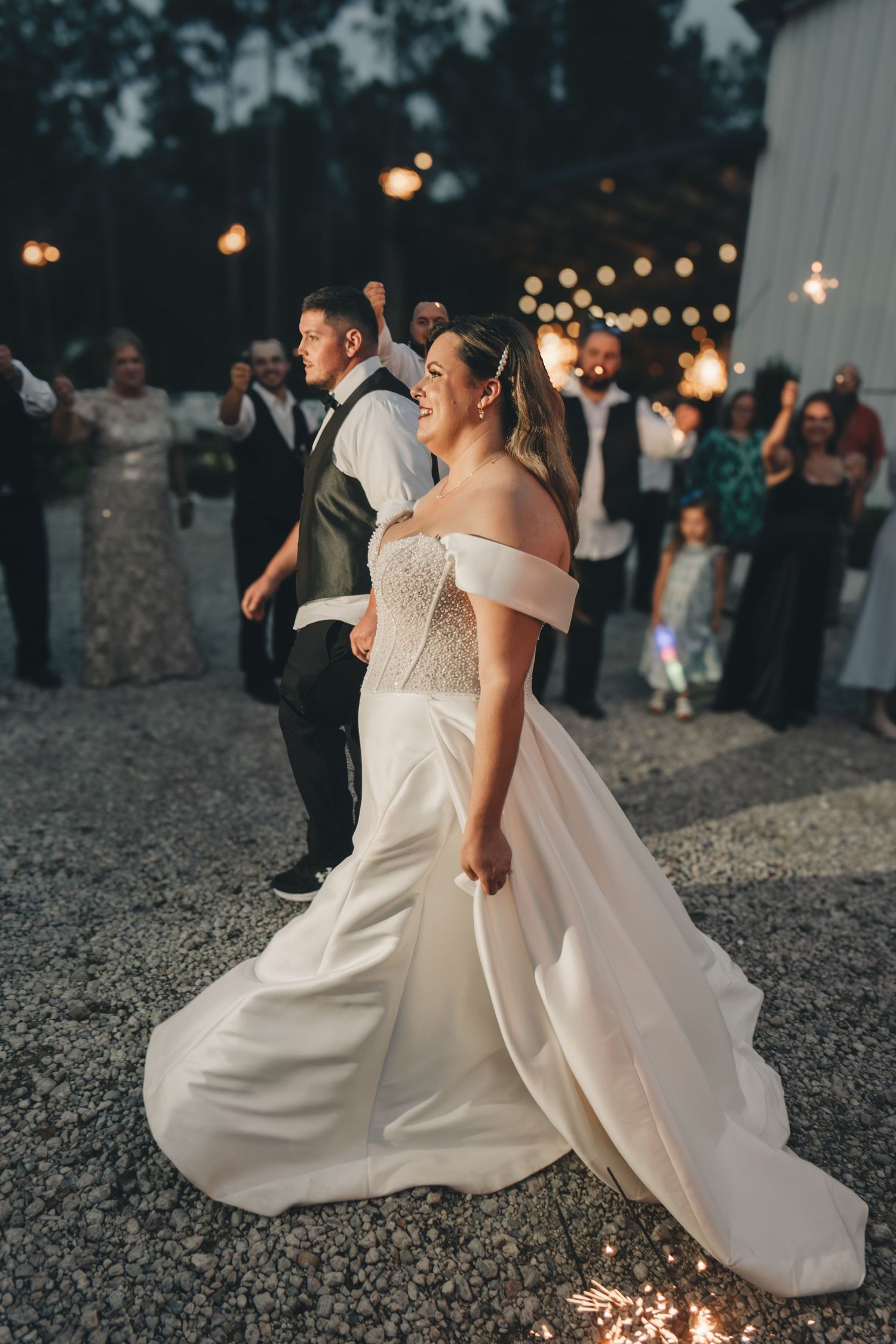 Bride in off-shoulder white gown smiles during sparkler exit at outdoor evening wedding reception.