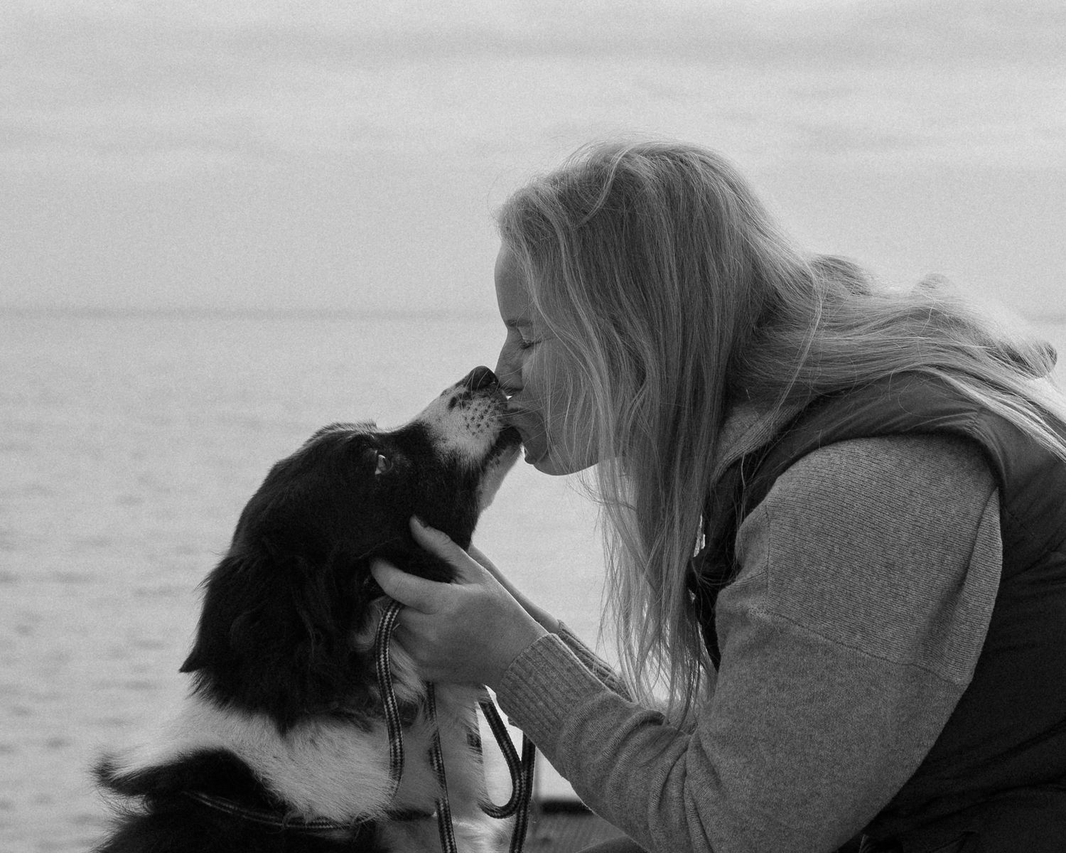 Black and white photo of a blonde woman kissing her black and white dog by the ocean.