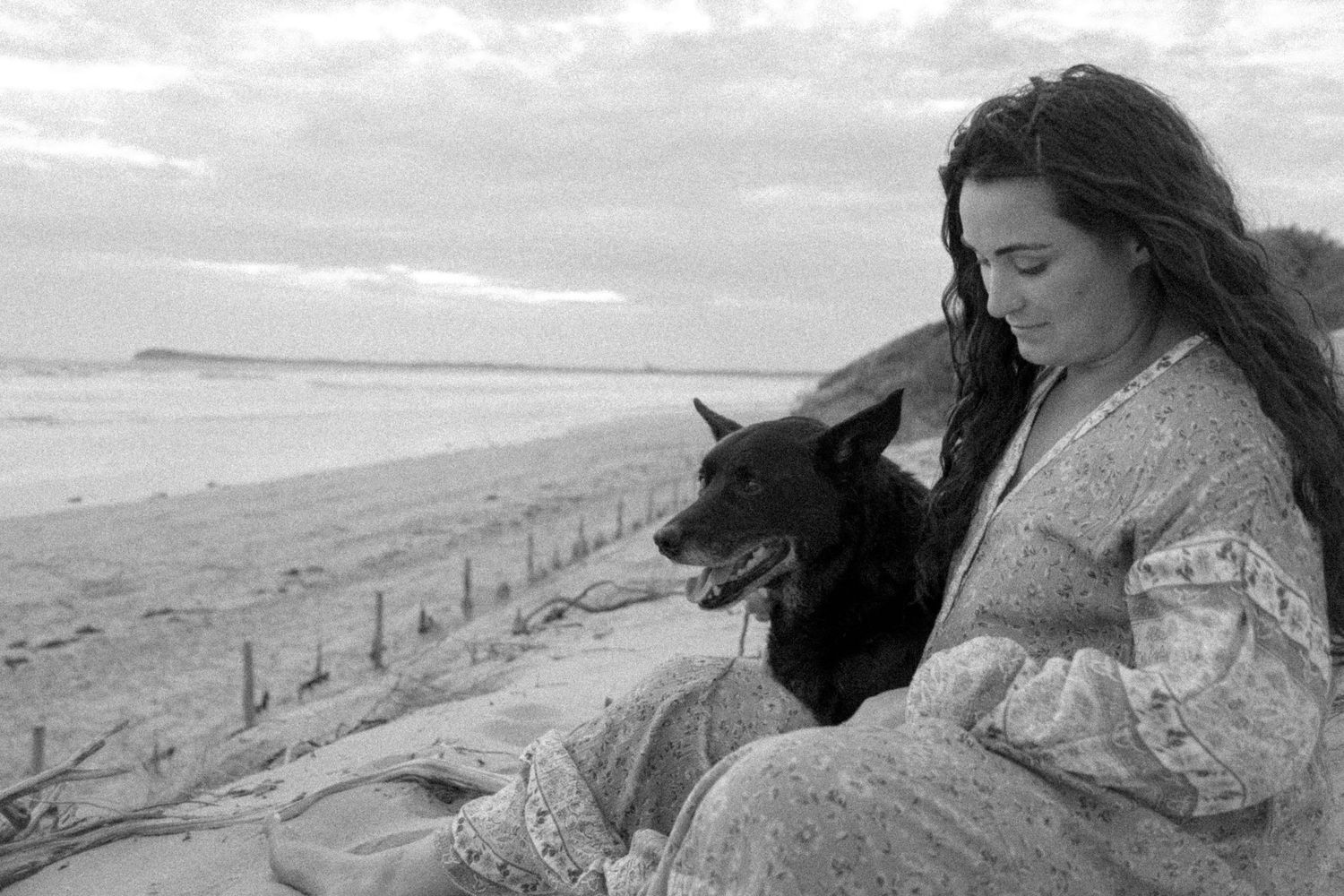 Woman sitting on a beach with a black dog in a serene black and white coastal scene.