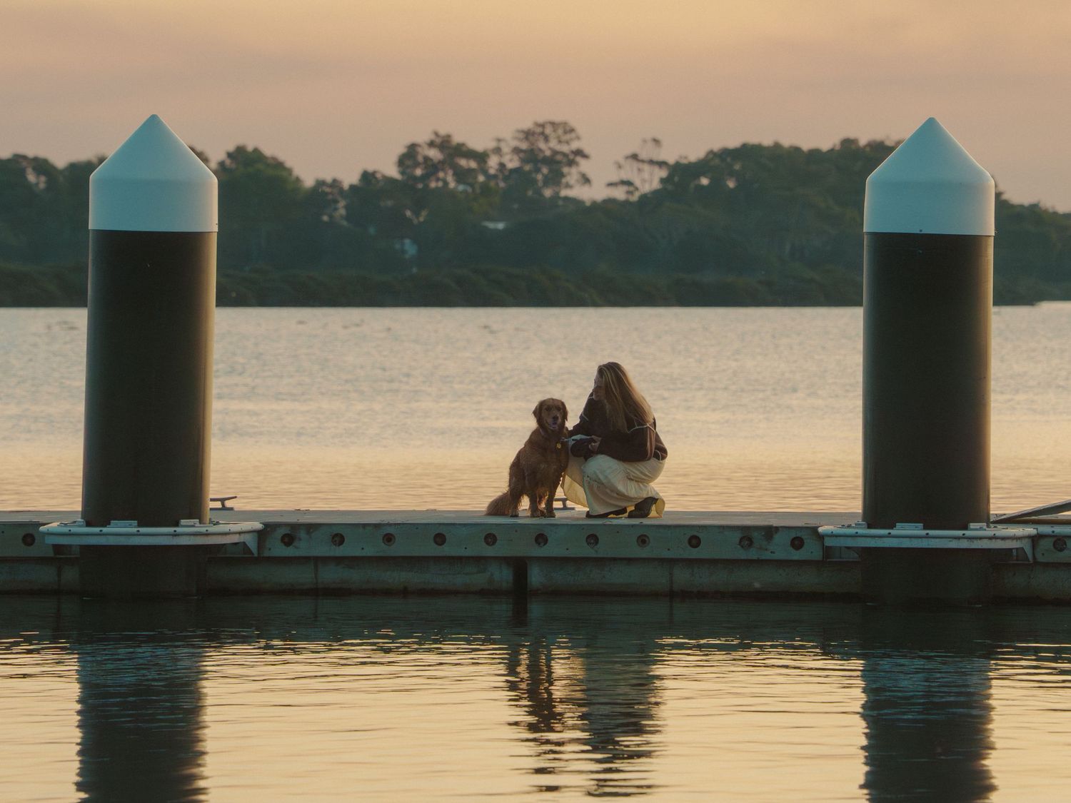 Woman and dog sitting together on a dock at sunset over calm water.