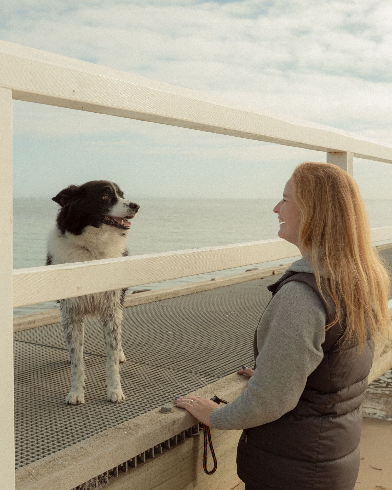 Blonde woman smiling at a black and white border collie standing on a seaside railing with ocean views.