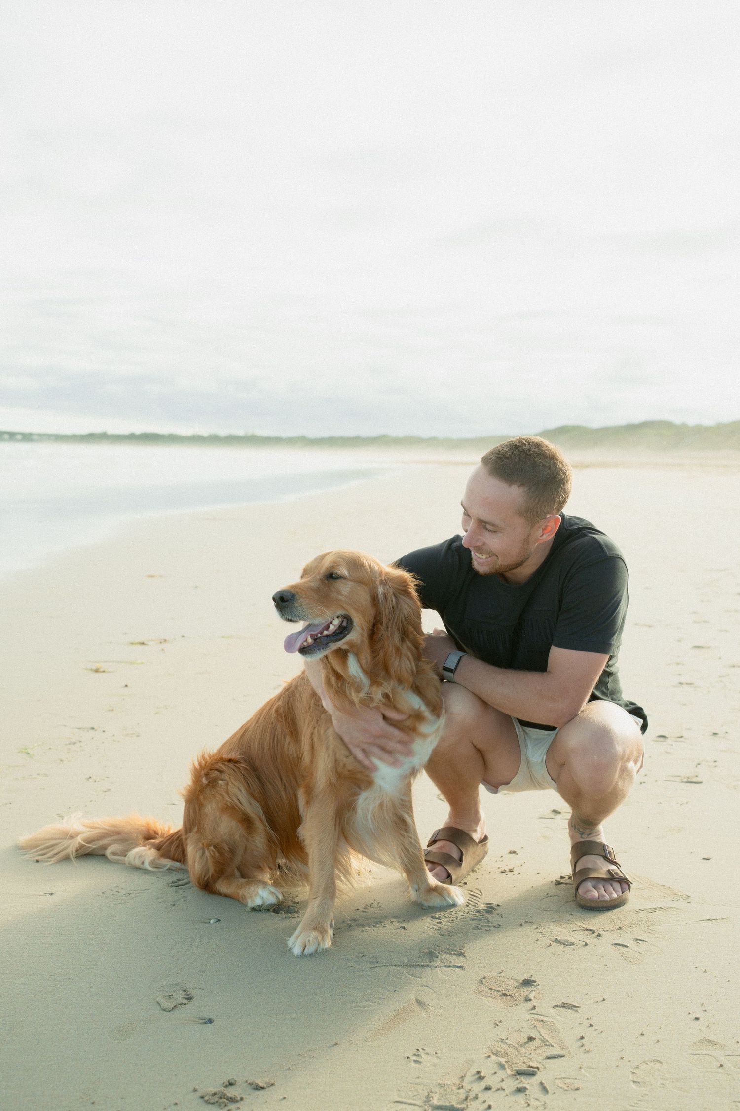 Man in black shirt crouching on sandy beach petting a golden retriever dog by the ocean.