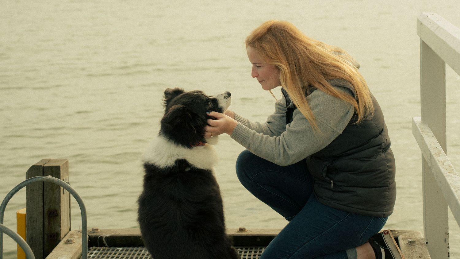 Blonde woman crouching on a pier interacting with a black and white border collie dog by the water.