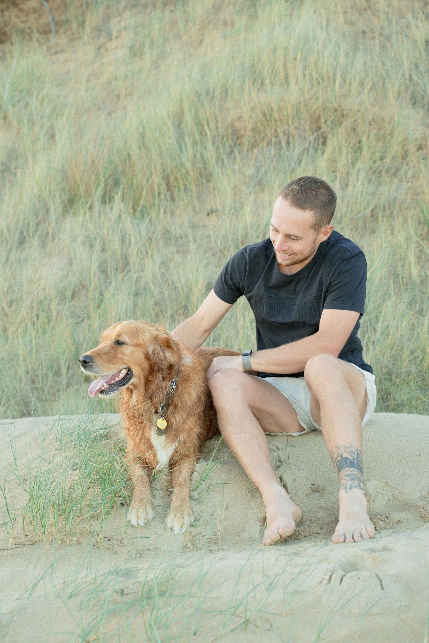 Man with tattoo sitting on sand dunes petting a golden retriever dog with grass in background.