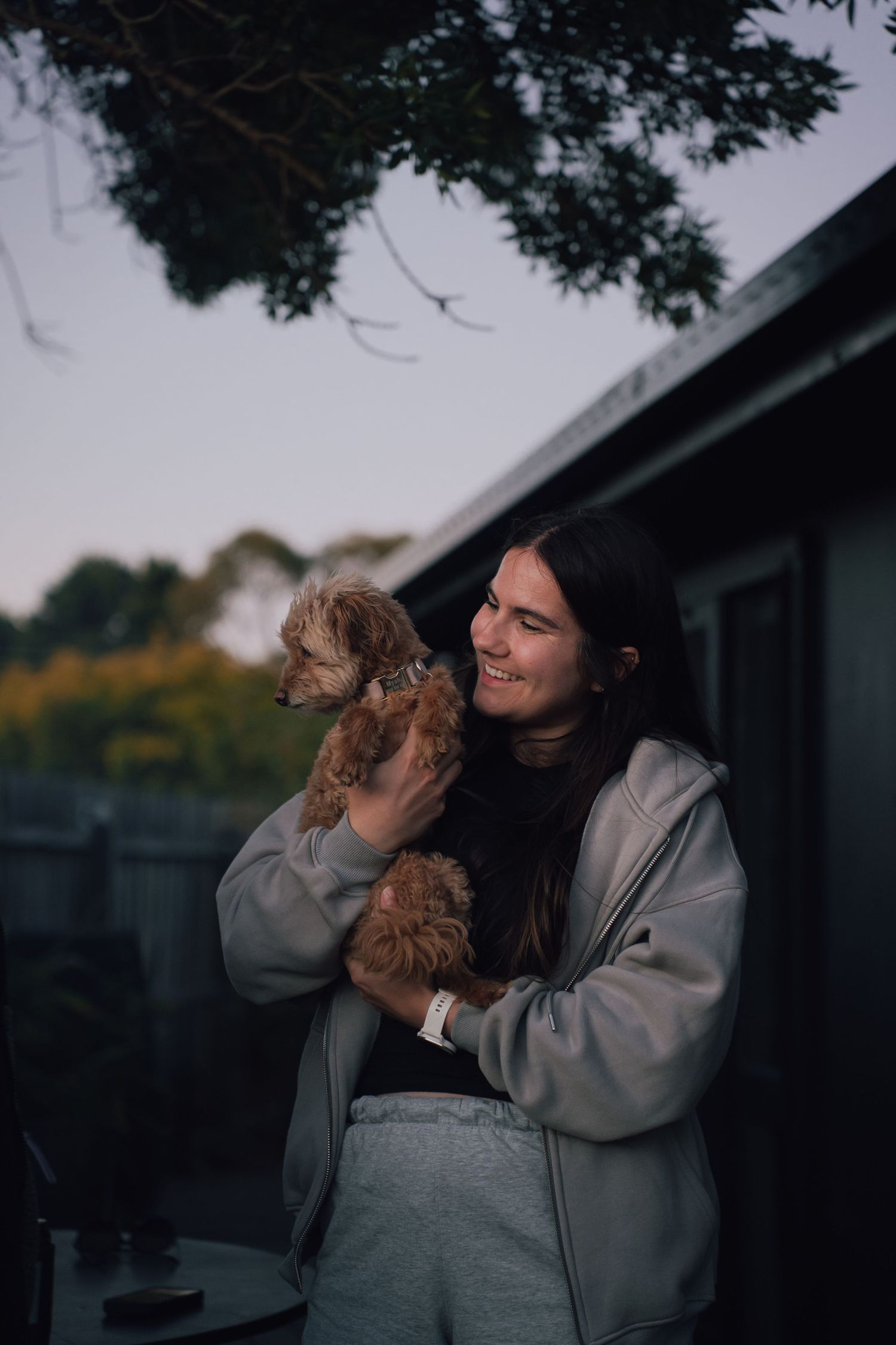 Young woman in grey hoodie smiling while holding a small fluffy golden cavoodle puppy outdoors at dusk.