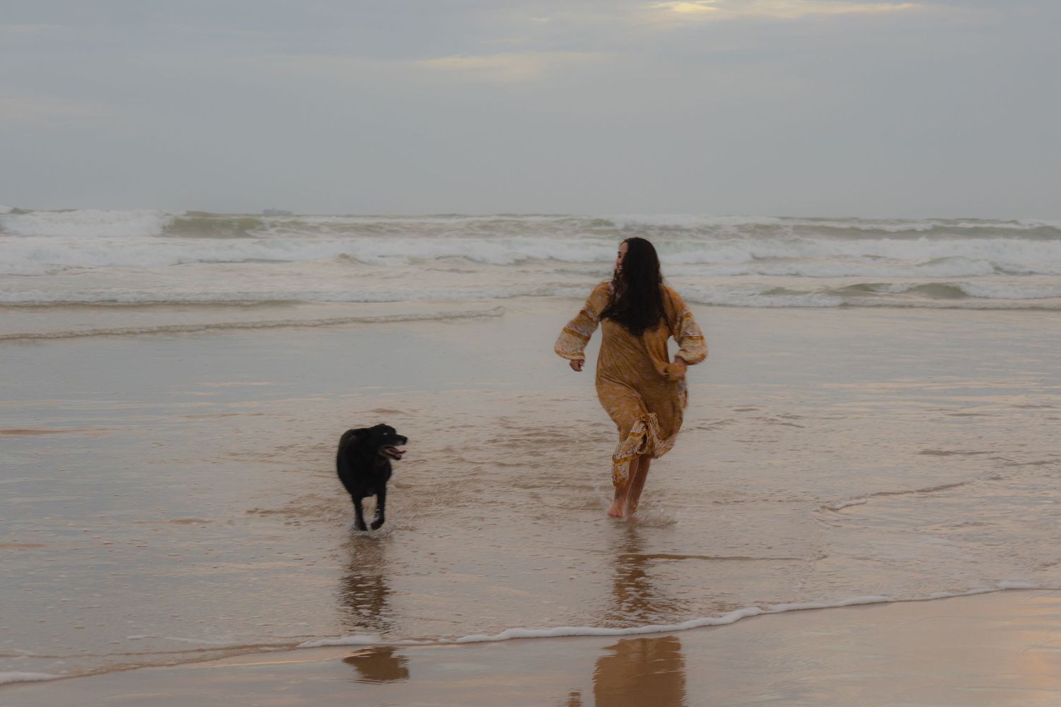 Woman in floral dress running with black dog along a beach at dusk.